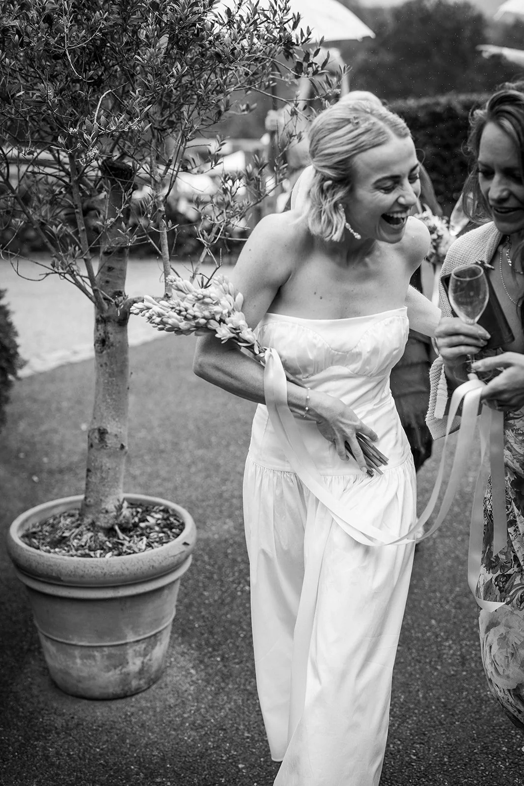A woman in a white strapless dress holding a bouquet of flowers and laughing with another woman at an outdoor event, with a potted tree and umbrellas in the background.