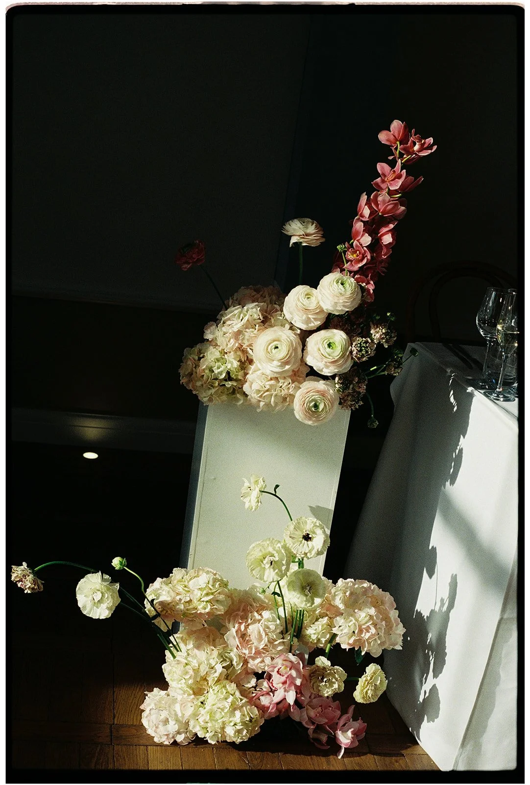 Arrangement of cream, pink, and white flowers in a white rectangular vase on a white table, with additional flowers in front, and glassware on the table to the right, set in a dimly lit room with a dark background.