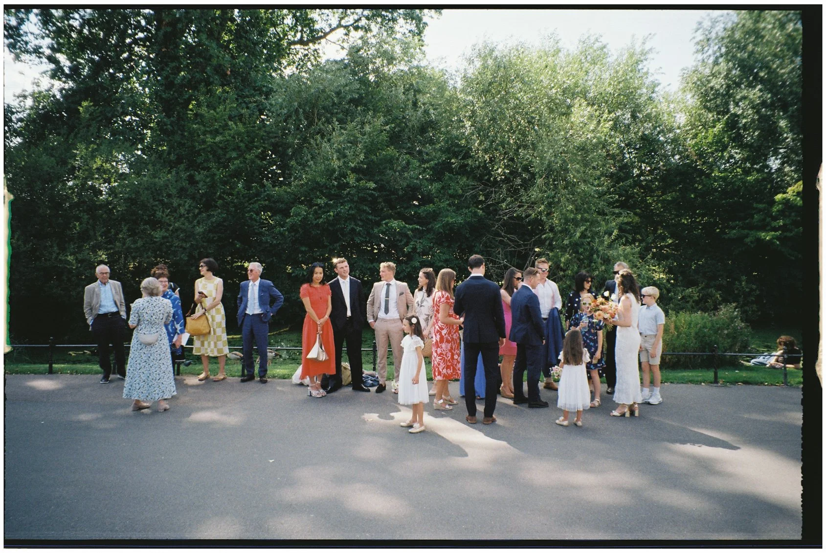 A group of people dressed in formal attire, including men, women, and children, gathered outdoors on a paved area with lush green trees in the background, possibly waiting for a wedding ceremony.
