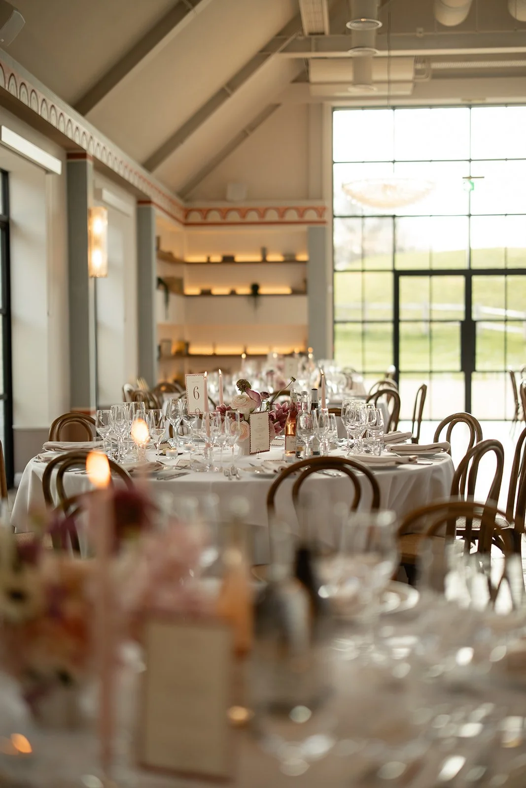 Inside a banquet hall decorated for a formal event with a round table set with multiple wine glasses, water bottles, floral centerpieces, and table numbers, illuminated by natural light from large windows.
