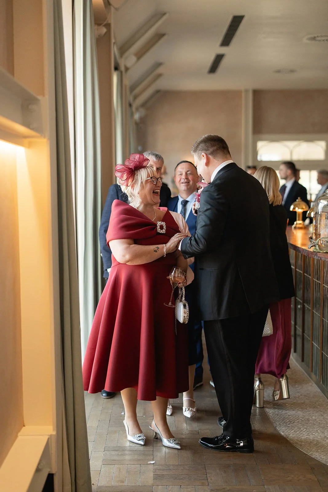 A woman in a red dress and matching hat talking to a man in a black suit at a social gathering, with other people in the background.