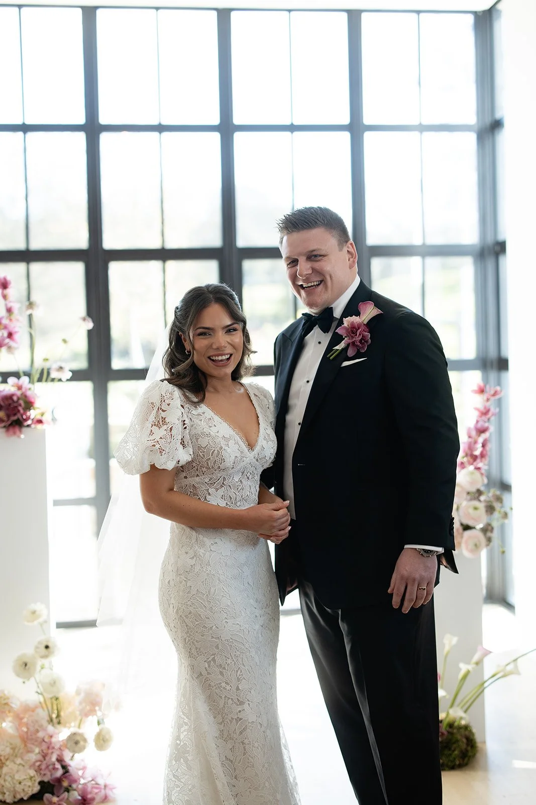 A happy couple in wedding attire standing inside a modern glass-walled venue, smiling and holding hands. The bride wears a lace gown with puff sleeves and a veil, while the groom wears a black tuxedo with a bow tie and purple flower boutonniere. Ther