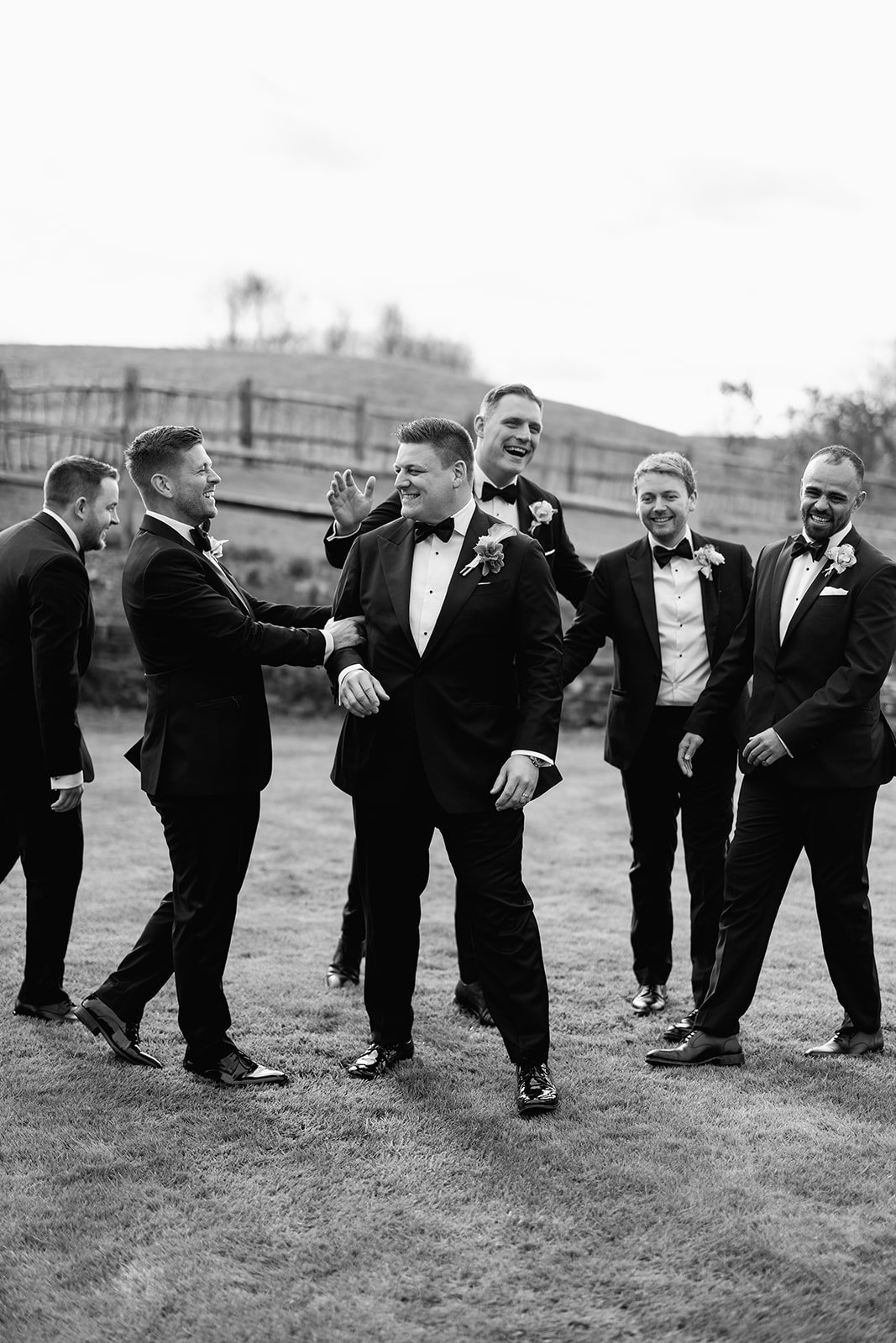 Black and white photo of six men in tuxedos with boutonnières, smiling and laughing outdoors on grass, with a wooden fence and hilly landscape in the background.