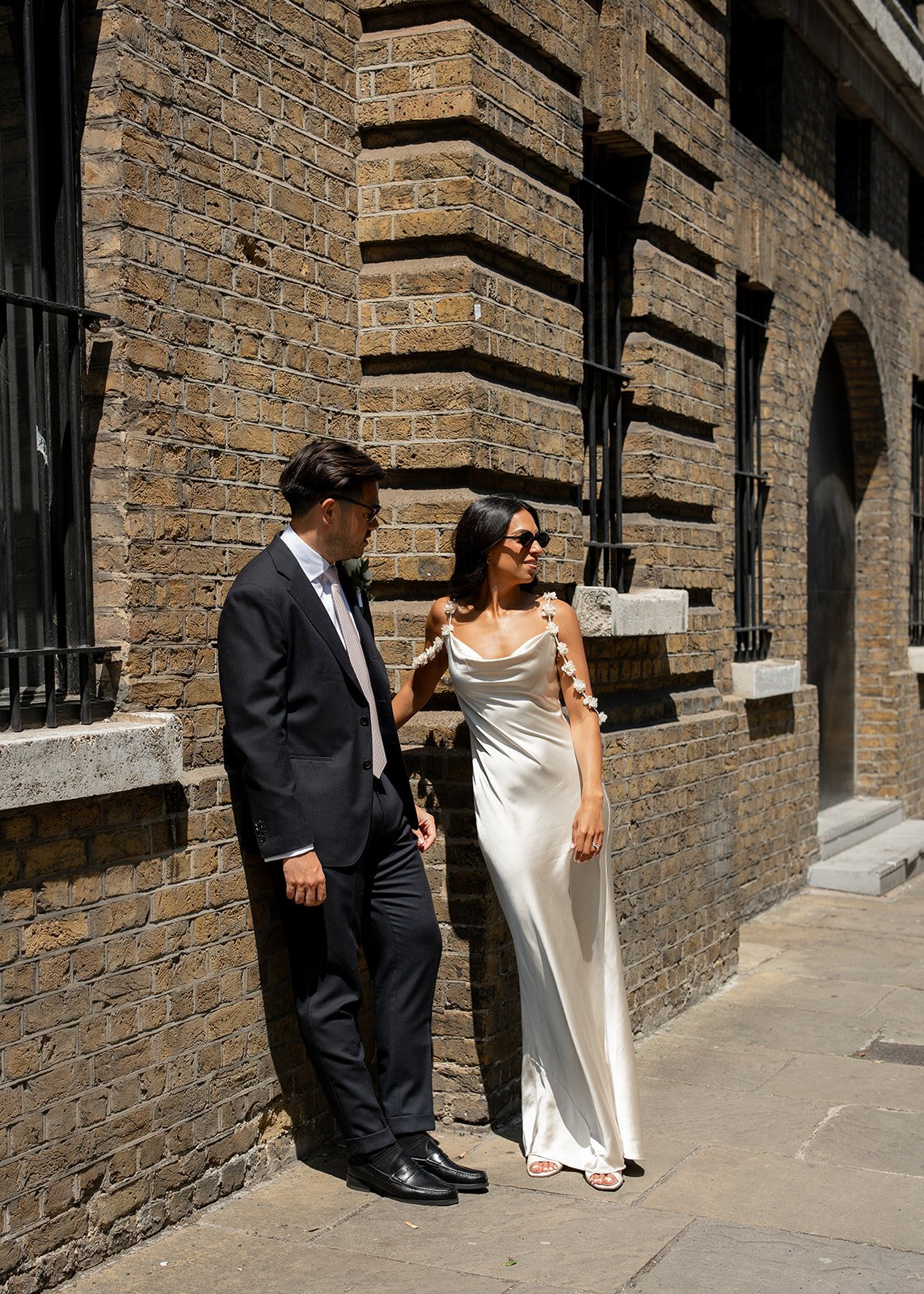 A man in a black suit and a woman in a white dress with floral shoulder details standing against a brick building. The woman is wearing sunglasses and smiling, while the man is looking down.