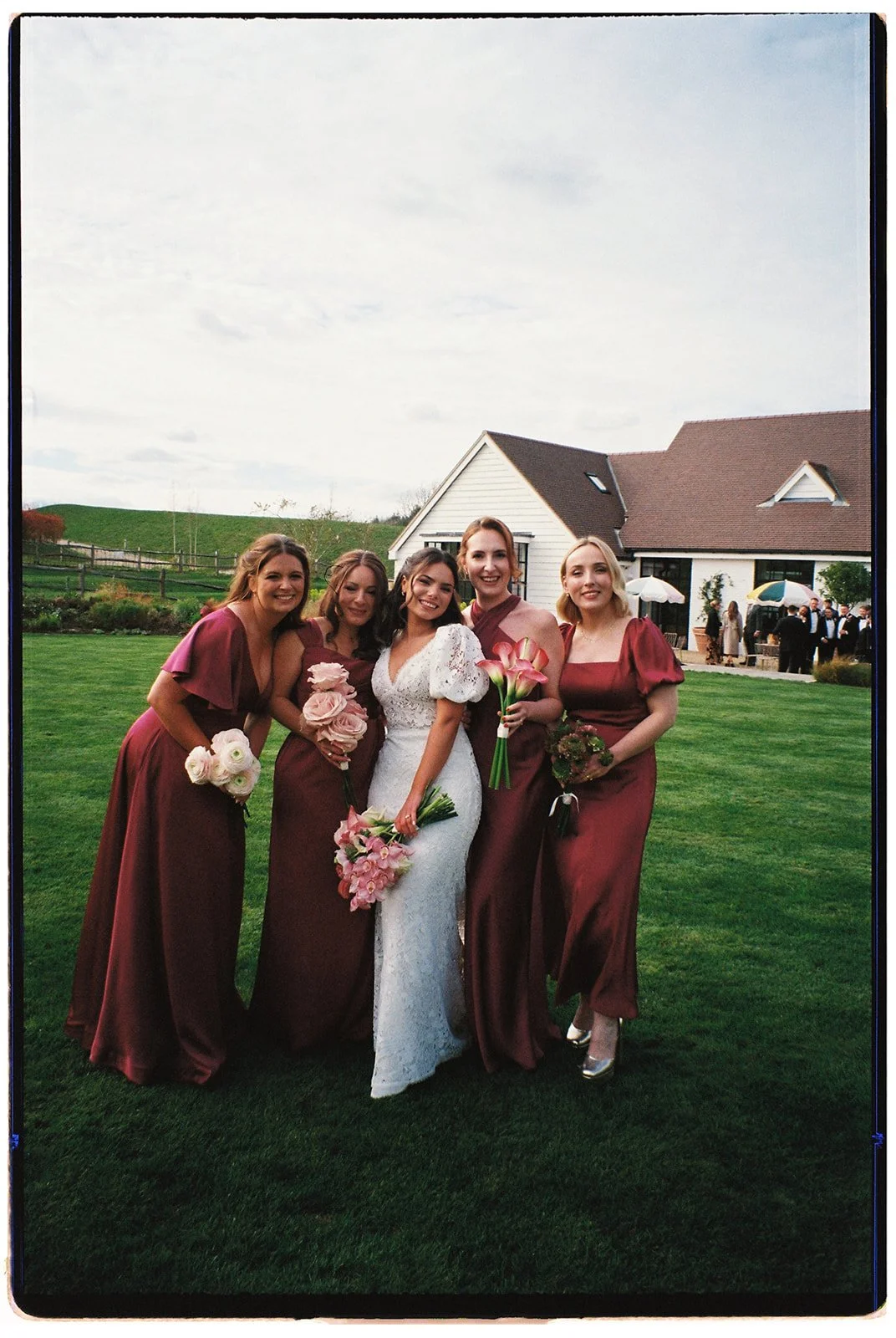A bride and five bridesmaids standing outdoors on a lawn, dressed in formal attire, holding bouquets of flowers, with a house and other guests in the background.