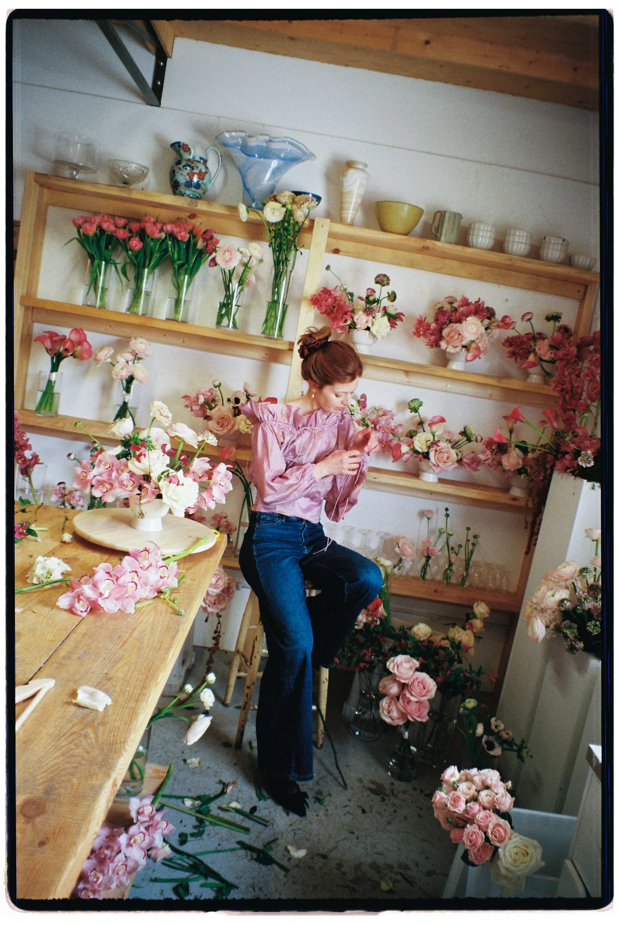A woman with a pink satin blouse and blue jeans sitting on a stool in a flower shop, arranging pink and white flowers with numerous floral arrangements and vases around her.