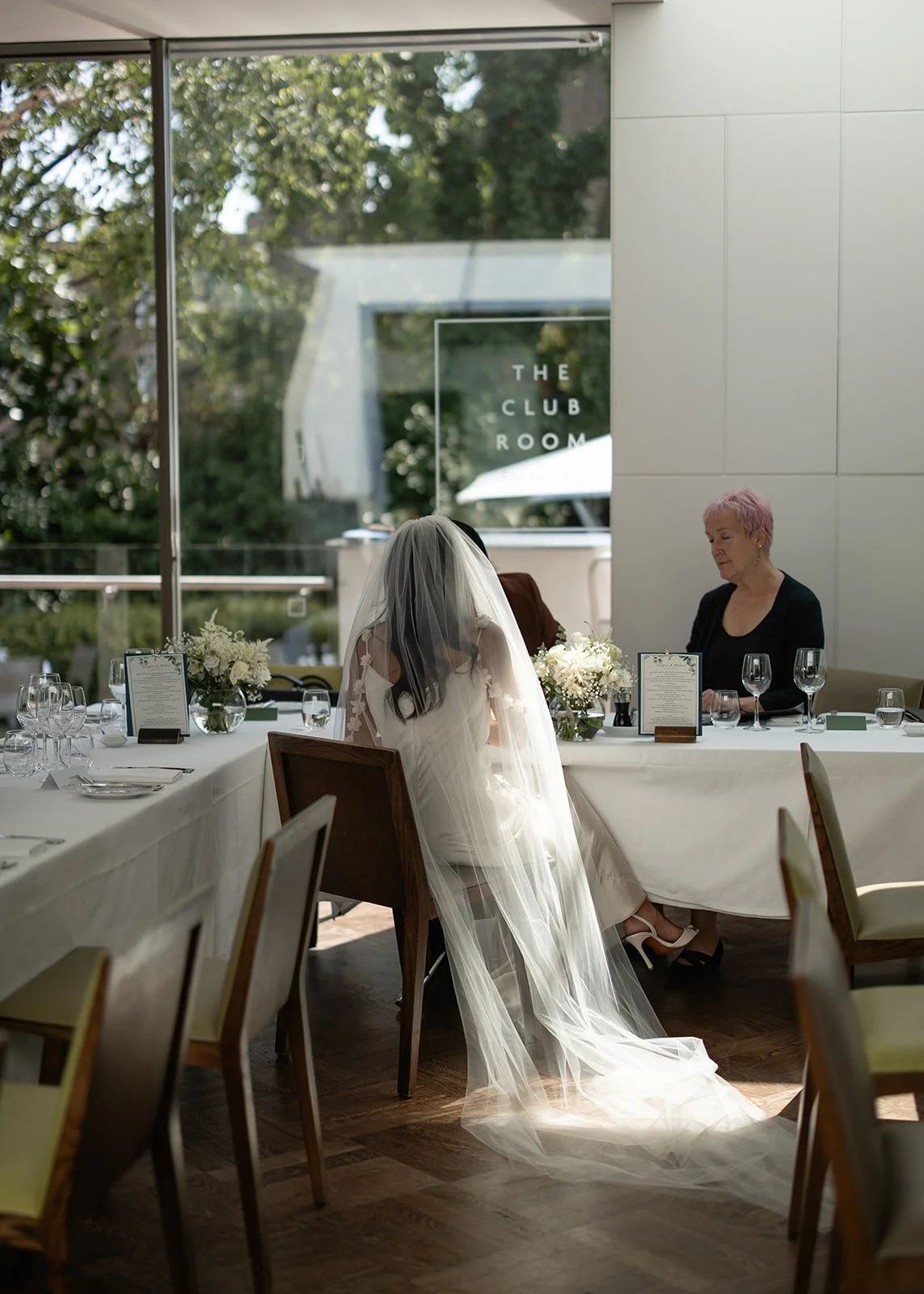 A bride in a wedding dress with a veil sitting at a dining table inside a restaurant, with a woman sitting across from her. The table is decorated with white flowers and set with glasses and silverware. Large windows in the background reveal greenery