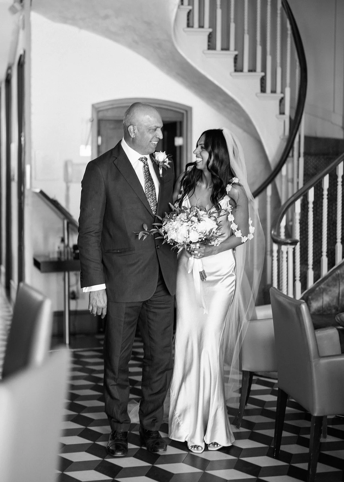 A bride and an older man are standing together inside a building with a staircase in the background, smiling at each other. The bride is holding a bouquet of flowers, wearing a wedding dress, and a veil. The man is dressed in a suit with a boutonnier