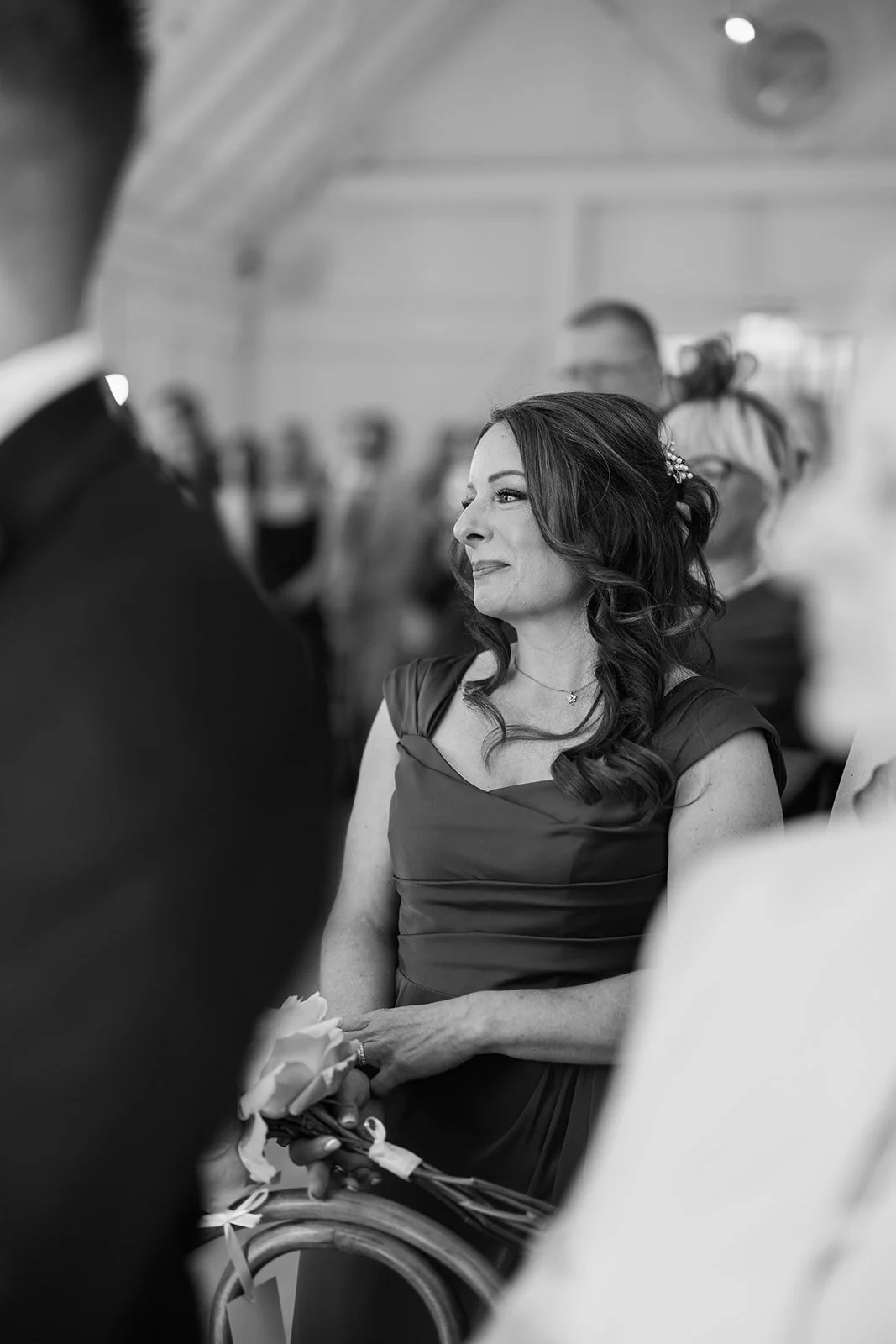 A woman with long dark hair and a dress, holding flowers, smiling and looking to the side, at a formal event with people in the background.