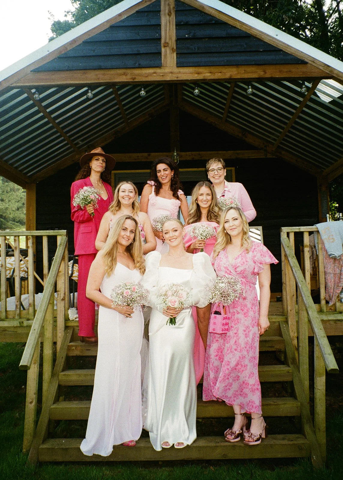 Group of women dressed in pink and white in a backyard, some holding flowers, posing on a wooden staircase in front of a covered porch.