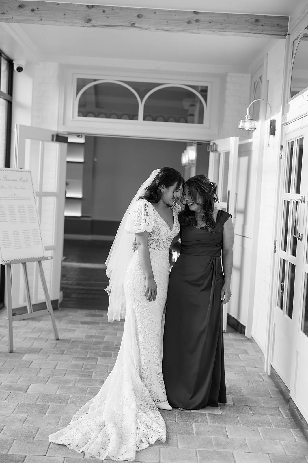 A bride in a lace wedding gown and veil sharing a joyful moment with a woman in a dark gown inside a well-lit indoor venue with white brick walls.