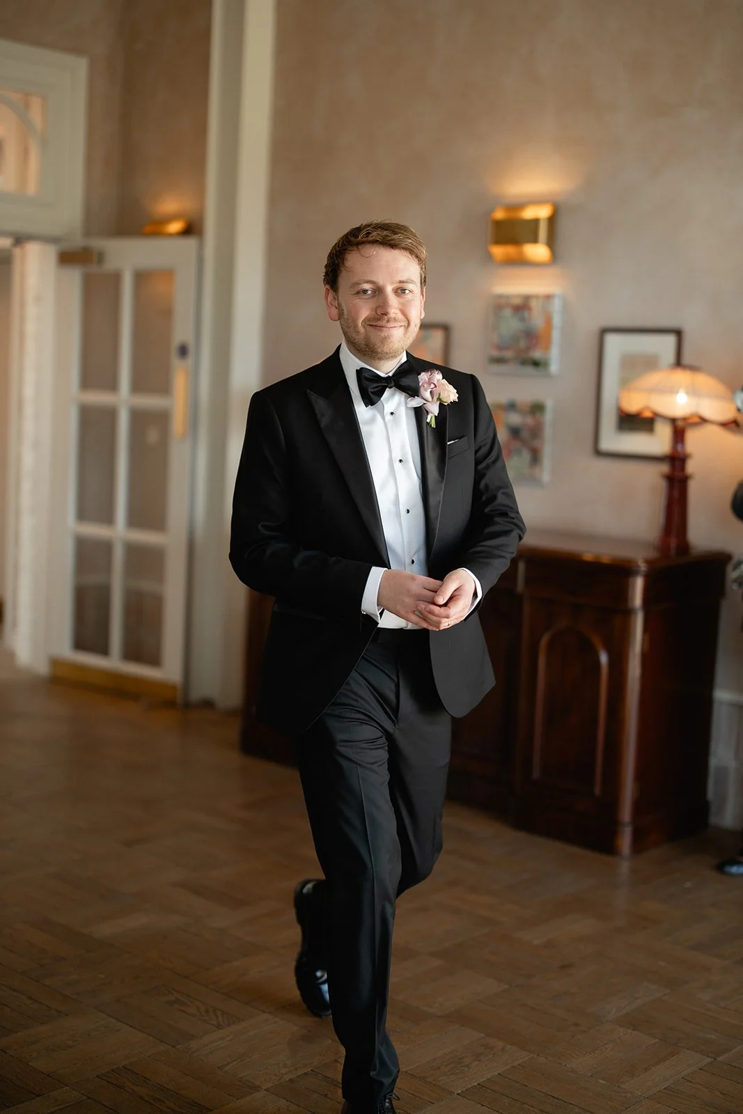 A man in a tuxedo with a bow tie and boutonniere walking inside a warmly lit room with wooden floors and framed artwork on the walls.