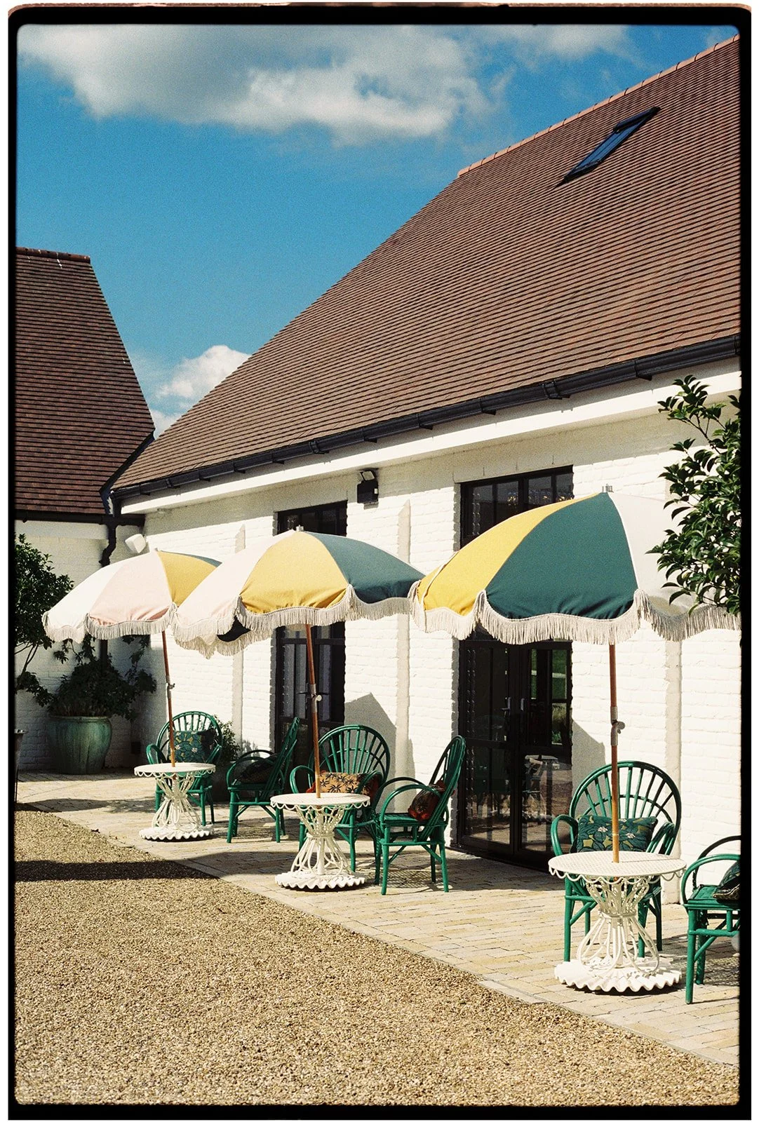 Outdoor patio area with green chairs, white tables, and multicolored umbrellas in front of a white brick building with brown shingled roof.