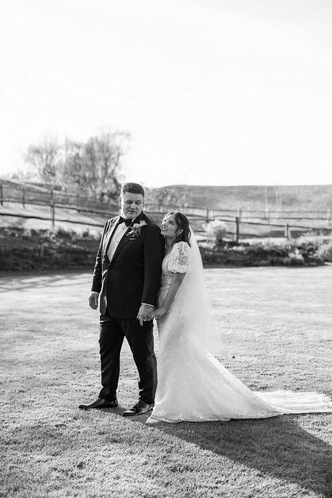 A black and white photograph of a bride and groom outdoors. The groom is wearing a tuxedo with a bow tie and boutonniere, and the bride is in a lace wedding gown with a long train. They are holding hands, with the bride smiling at the groom. In the b