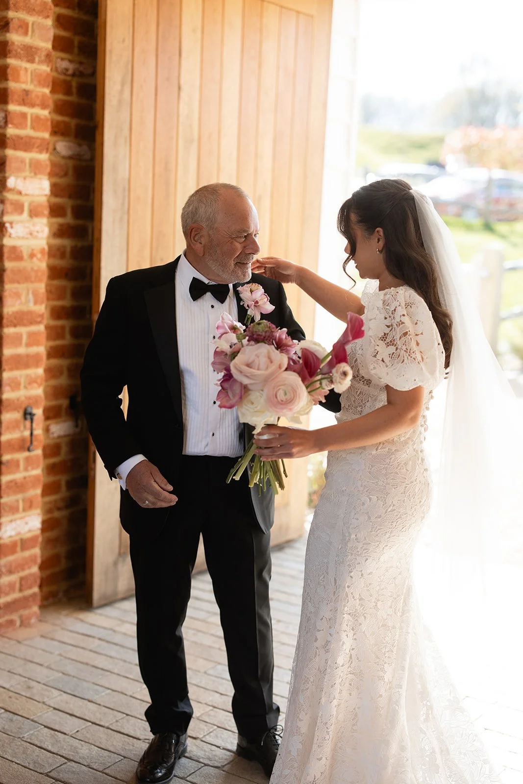 A bride in a lace wedding dress and a veil is giving a bouquet of pink and white flowers to an older man in a tuxedo, inside a rustic venue with brick and wood walls.