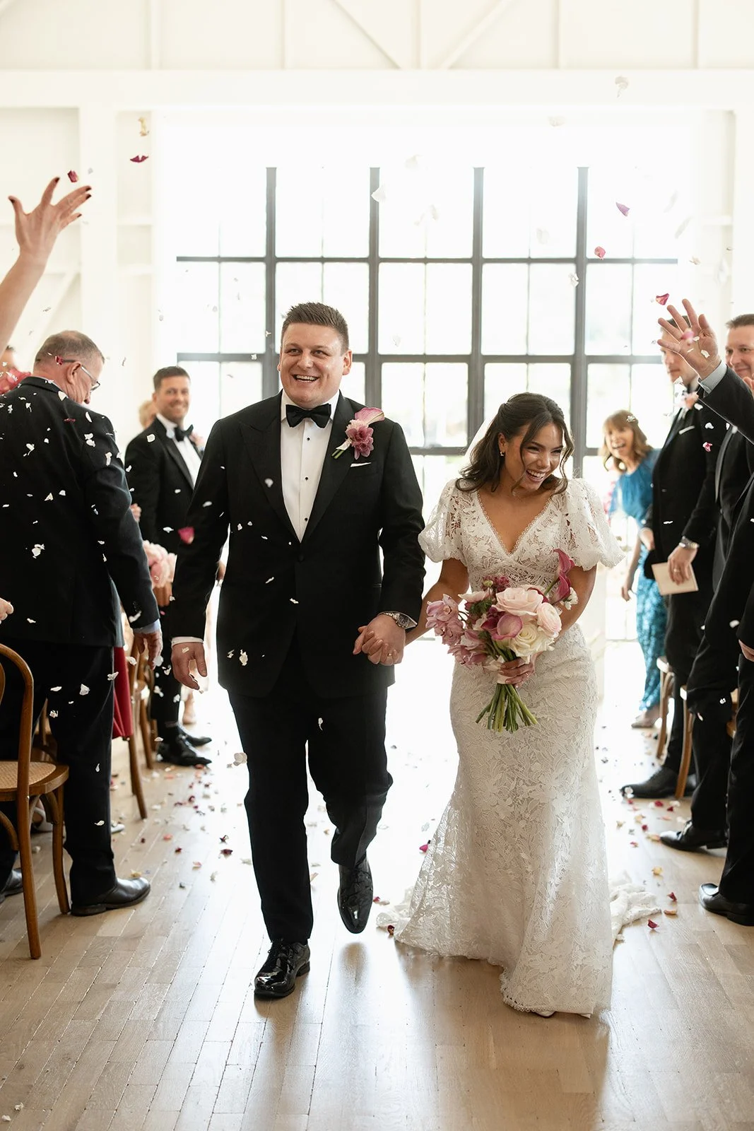 A bride and groom walking hand-in-hand down an aisle during their wedding ceremony, smiling, with guests throwing flower petals.