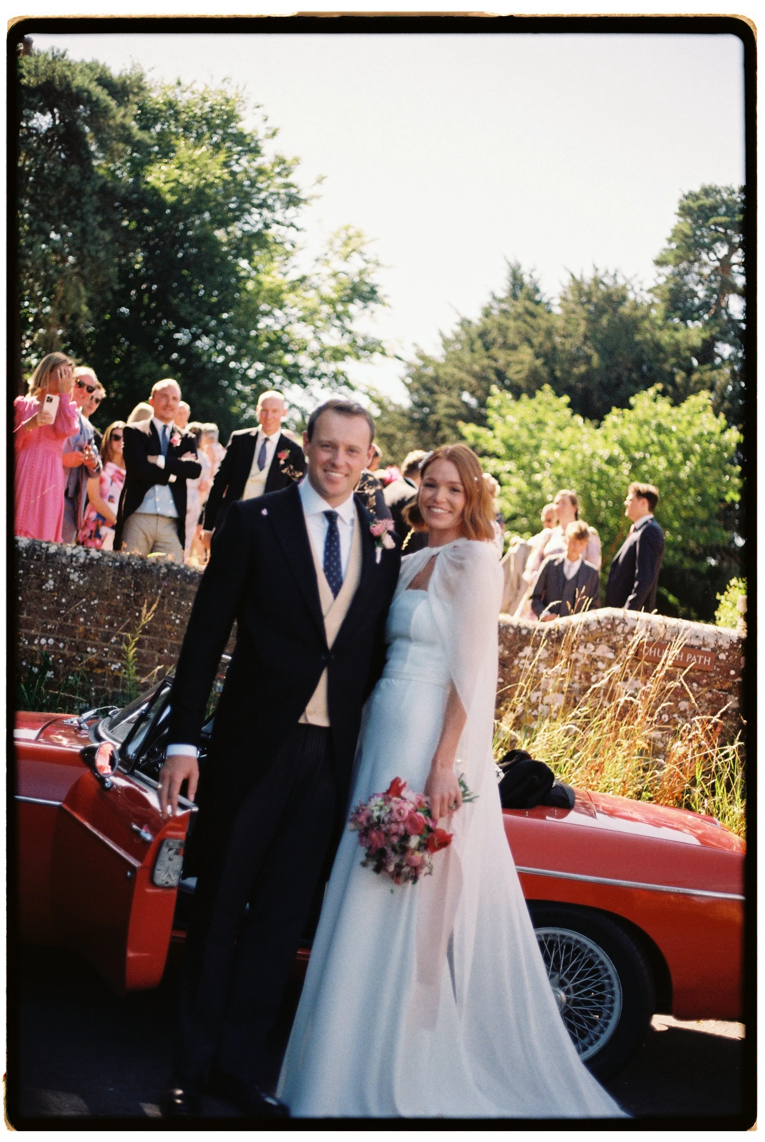 A newlywed couple standing in front of a red vintage convertible car, with wedding guests behind them on a brick wall, under bright daylight and green trees.