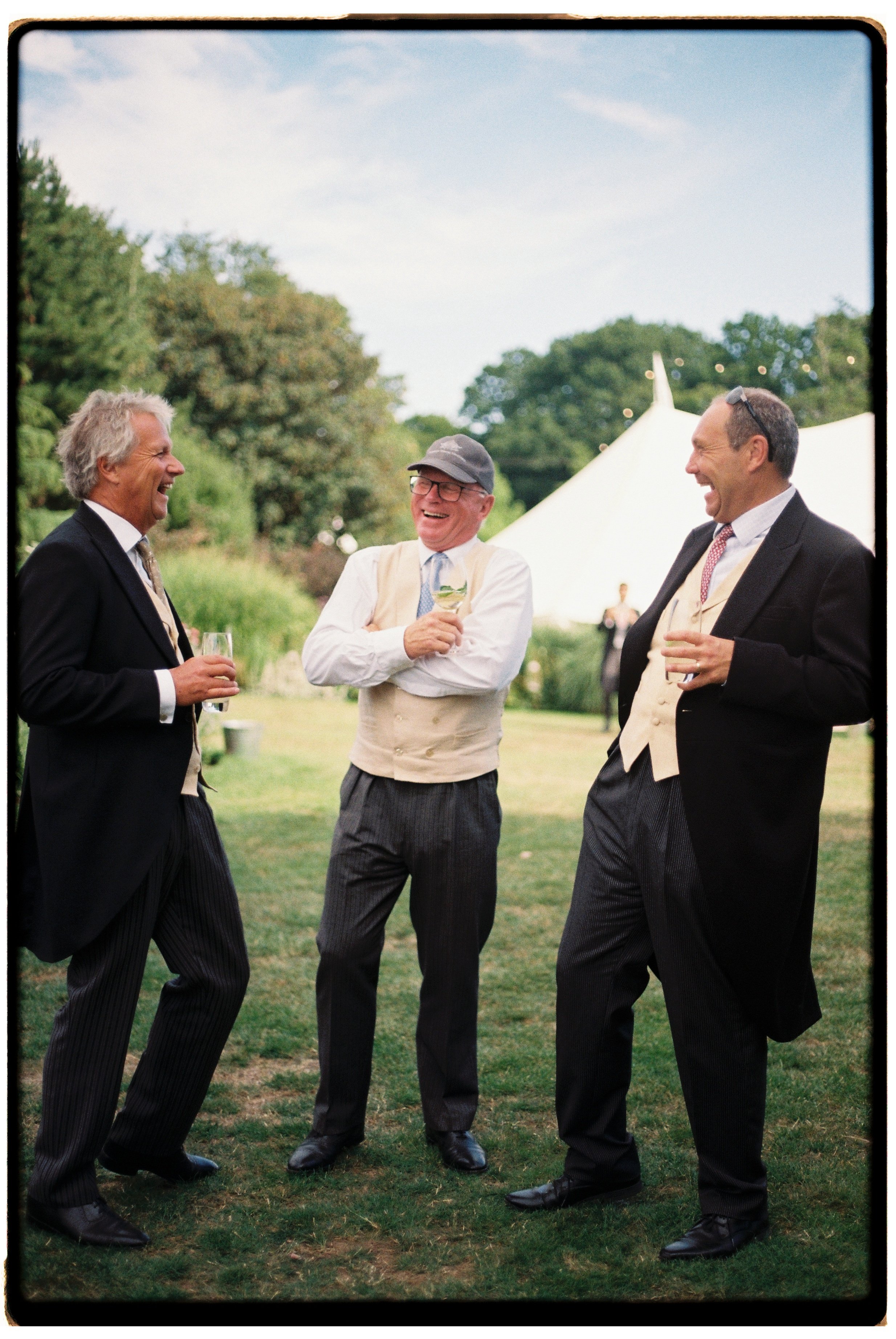 Three men in formal wear laughing and enjoying drinks outdoors at a garden party or wedding reception.