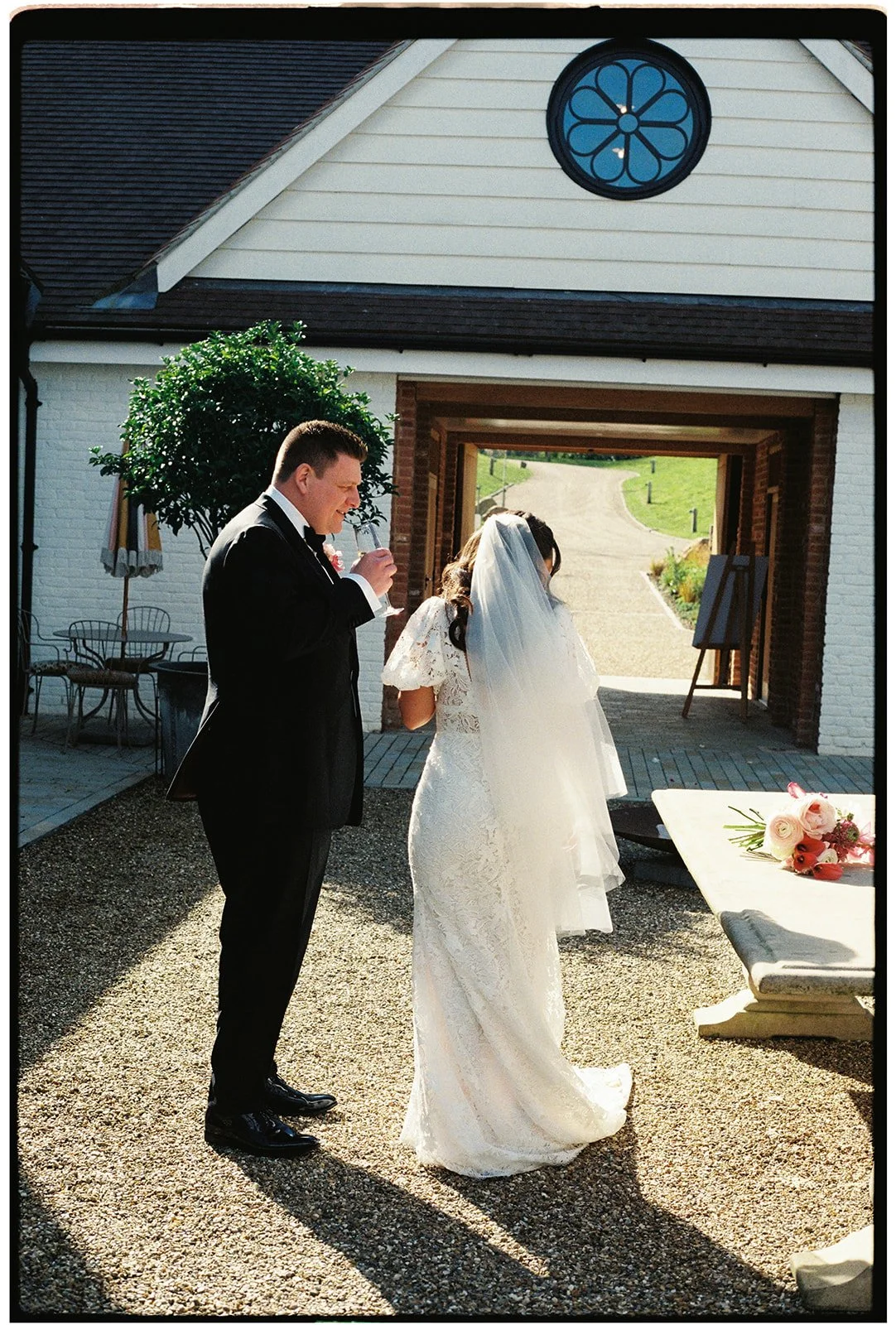 A bride and groom standing outside a building, with the groom drinking from a glass and the bride wearing a veil, during a wedding ceremony or celebration.