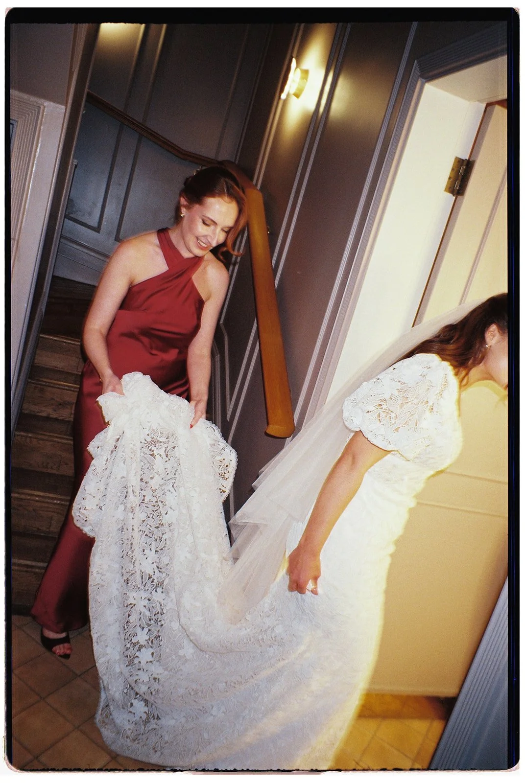 A woman in a burgundy satin dress is holding a white lace wedding veil, standing near a staircase in a home. Another woman, partially visible, is wearing a white wedding gown and veil, adjusting her dress.