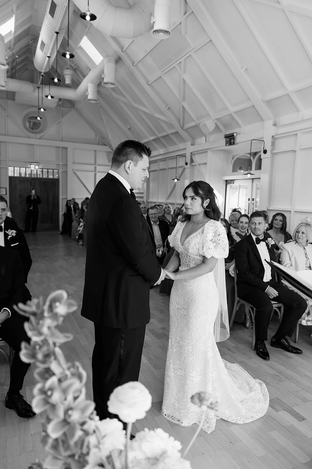 Black and white photo of a bride and groom holding hands during a wedding ceremony inside a bright, modern venue with wooden floors and a vaulted ceiling. Guests are seated in the background, watching the ceremony.