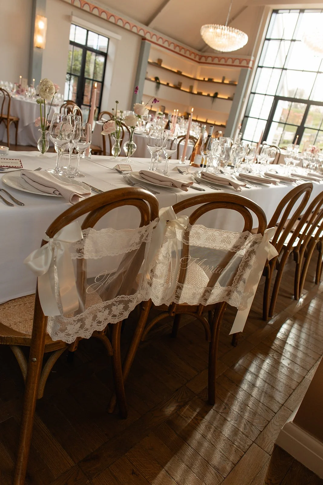 Elegant wedding reception setup with round and rectangular tables, white tablecloths, floral centerpieces, glassware, and candles, in a bright room with large windows and a chandelier.
