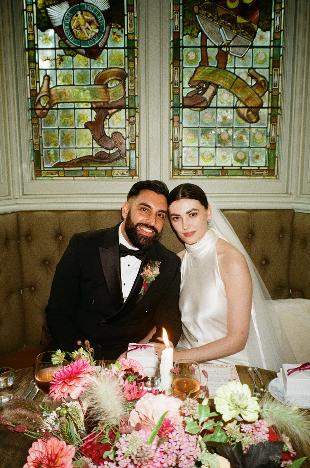 A newly married couple sitting at a table with a floral centerpiece and a lit candle, smiling, with stained glass windows in the background.