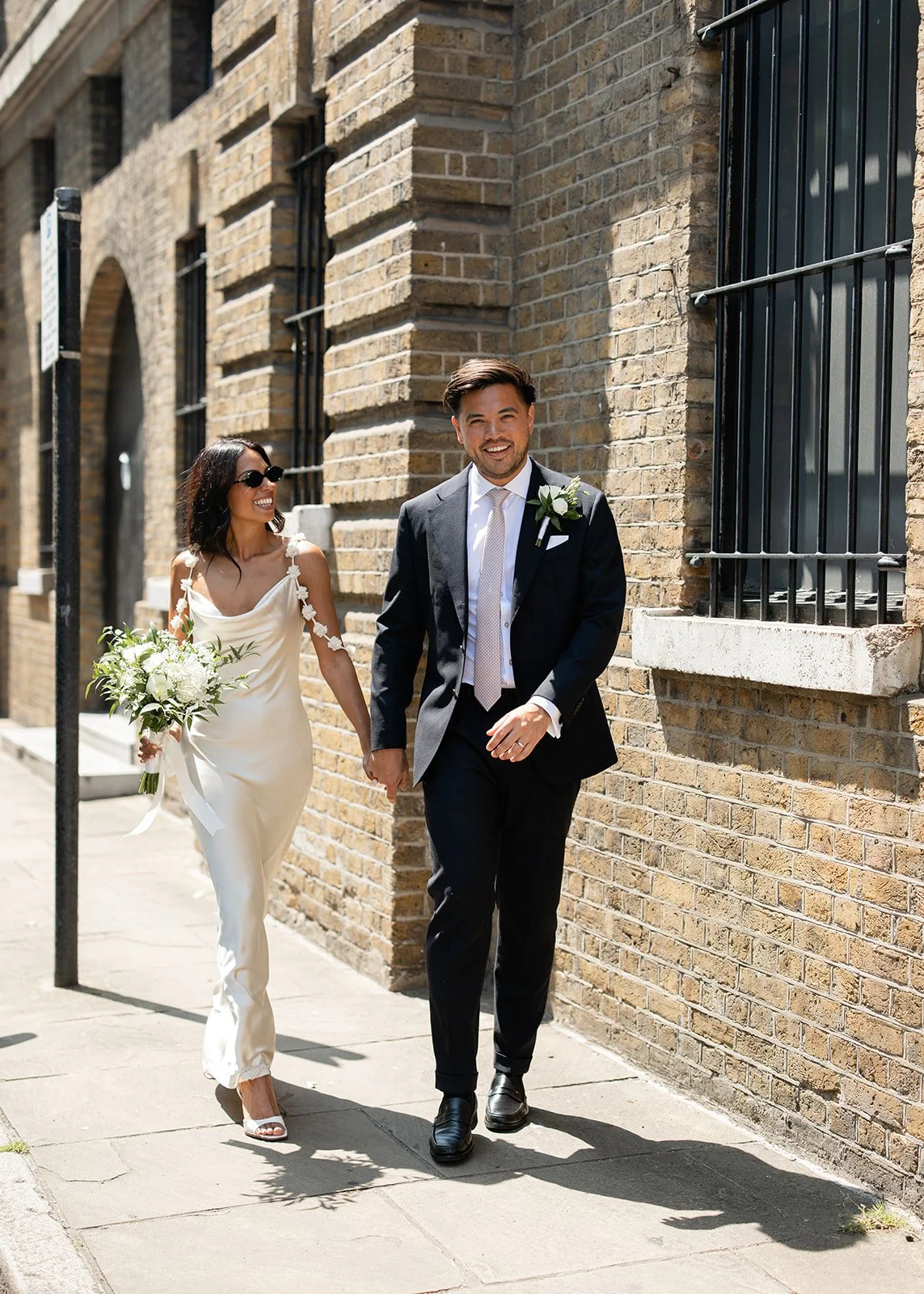 A happy couple walking hand in hand on a city sidewalk during their wedding day. The woman is wearing a white wedding dress and sunglasses, holding a bouquet of white flowers. The man is dressed in a dark suit with a white shirt and tie, with a bouto