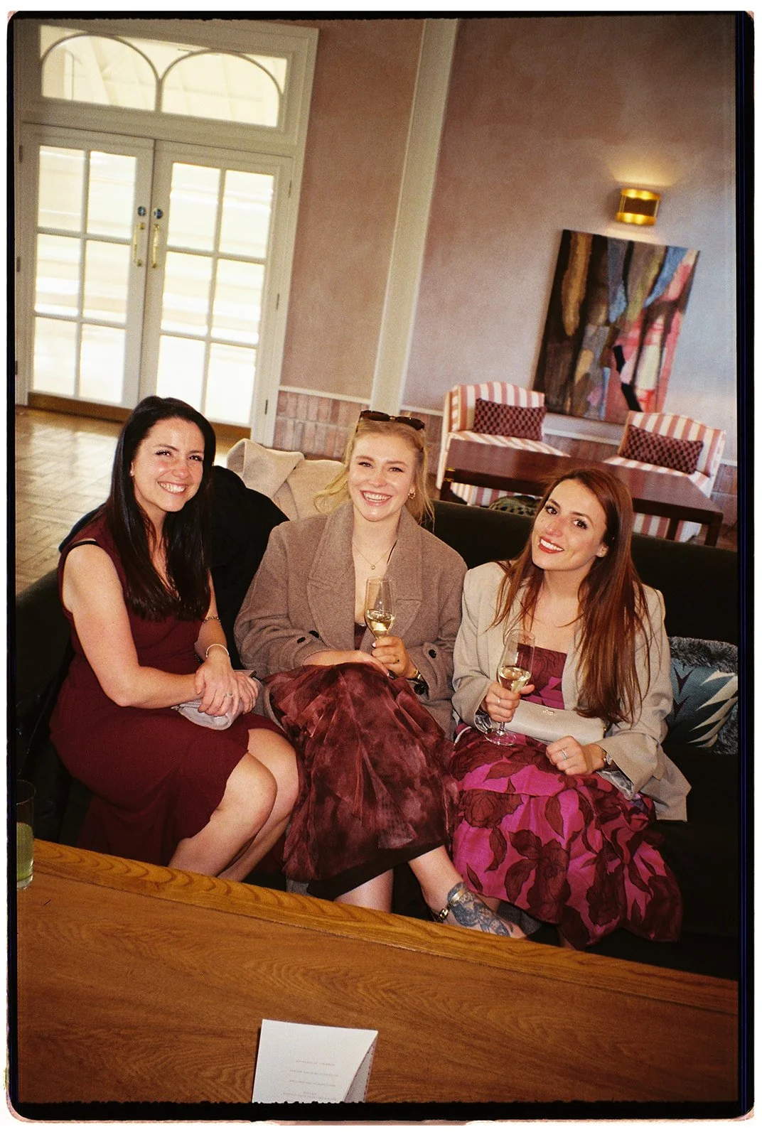 Three women sitting on a sofa, holding wine glasses, smiling at the camera in a well-lit room with a door and colorful artwork behind them.