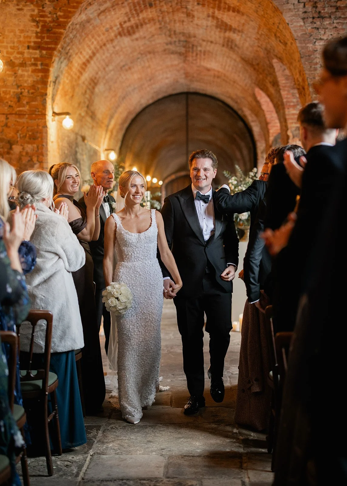 Bride and groom walking down the aisle at their wedding ceremony in a brick-walled venue, surrounded by guests clapping and smiling.
