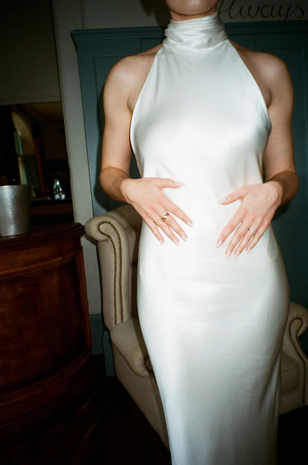 A woman in a silky white dress with a high collar, standing indoors, with her hands resting on her waist, wearing rings on her fingers.