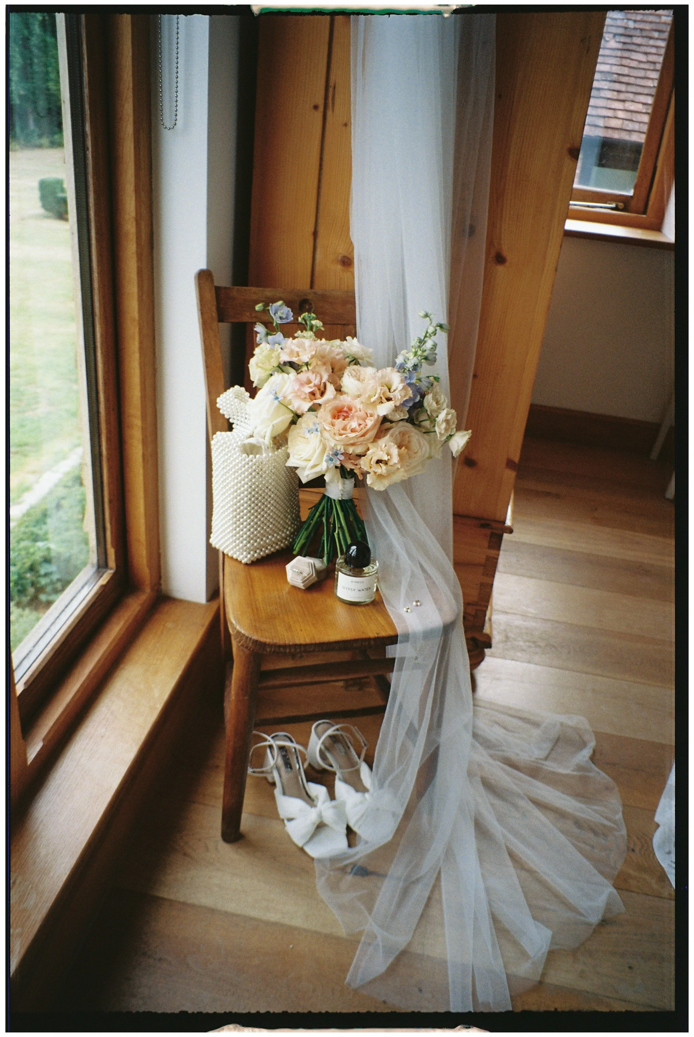A wooden chair next to a window with a bouquet of flowers, a white textured handbag, a small perfume bottle, a pair of white high-heeled shoes with bows, and a sheer white fabric draped over the chair.