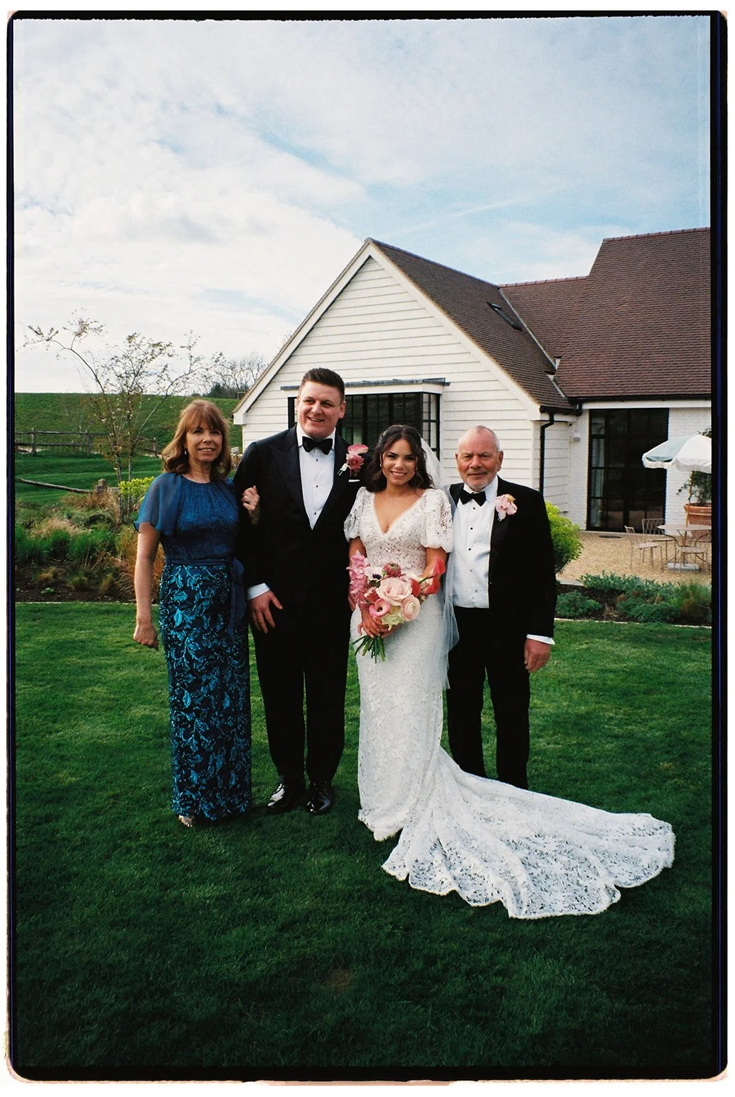 A bride and groom with two family members standing outdoors in front of a white house with a brown roof, on a grassy lawn during a wedding celebration. The bride is holding a bouquet of pink and white flowers, and both the bride and groom are dressed