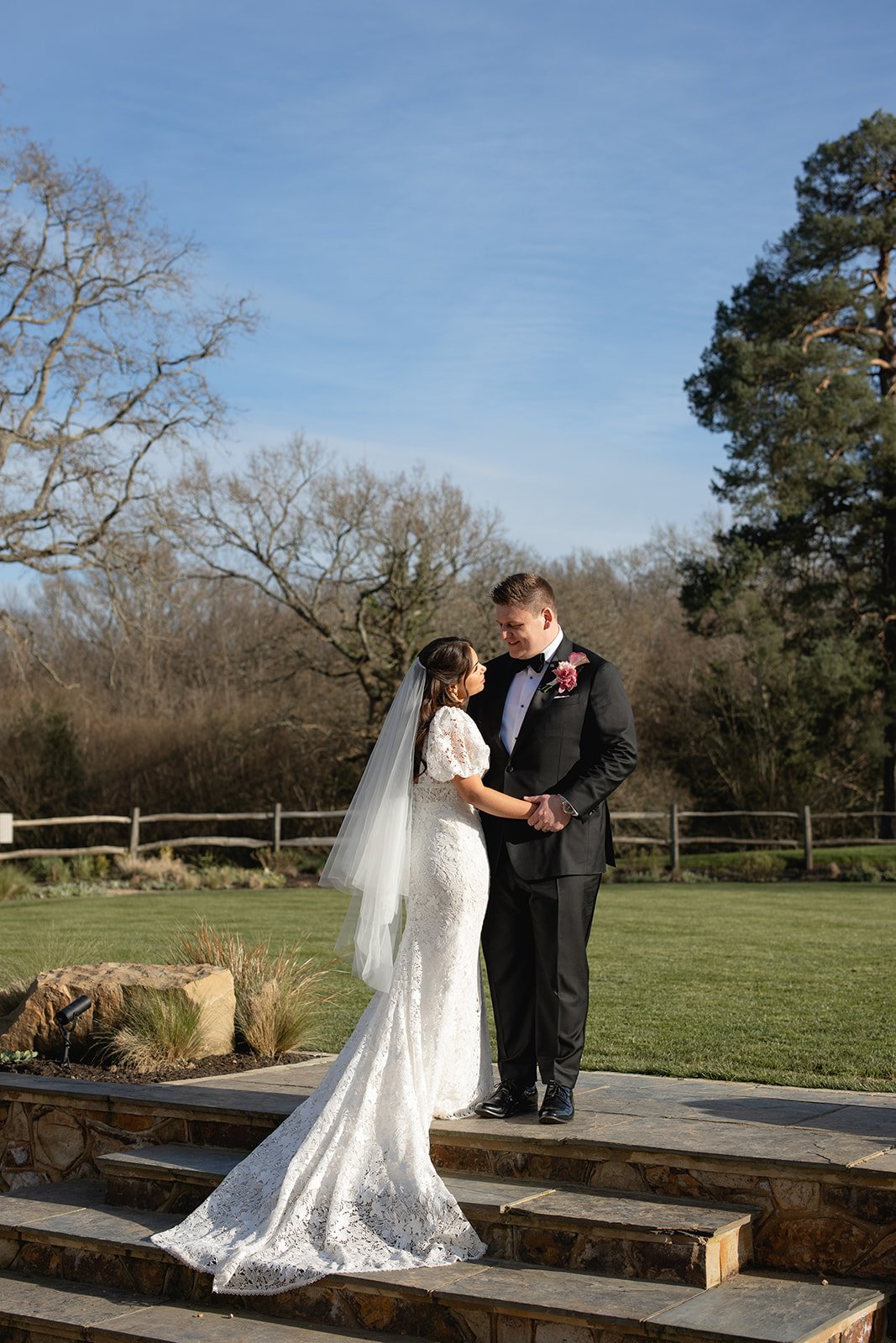 A bride and groom holding hands and looking at each other, standing on steps outdoors during a wedding ceremony, with trees and a grassy garden in the background on a clear day.
