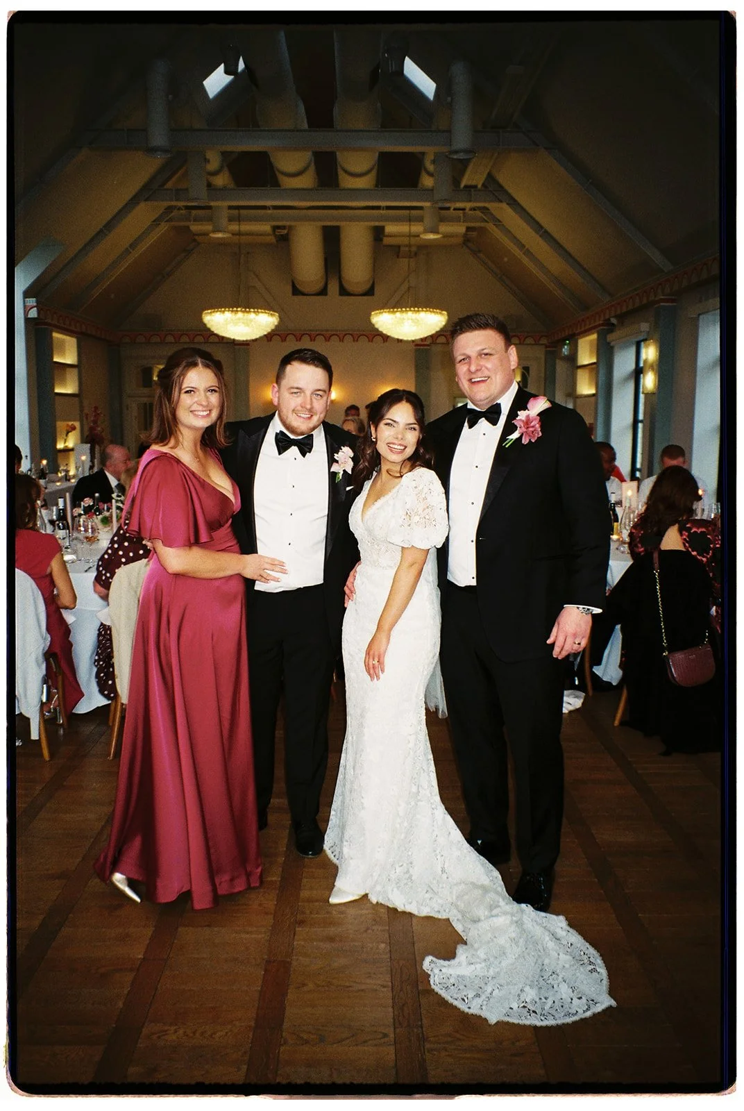 Group of four people at a wedding reception, including a bride in a white wedding dress, two men in tuxedos, and a woman in a burgundy gown, inside a decorated banquet hall.