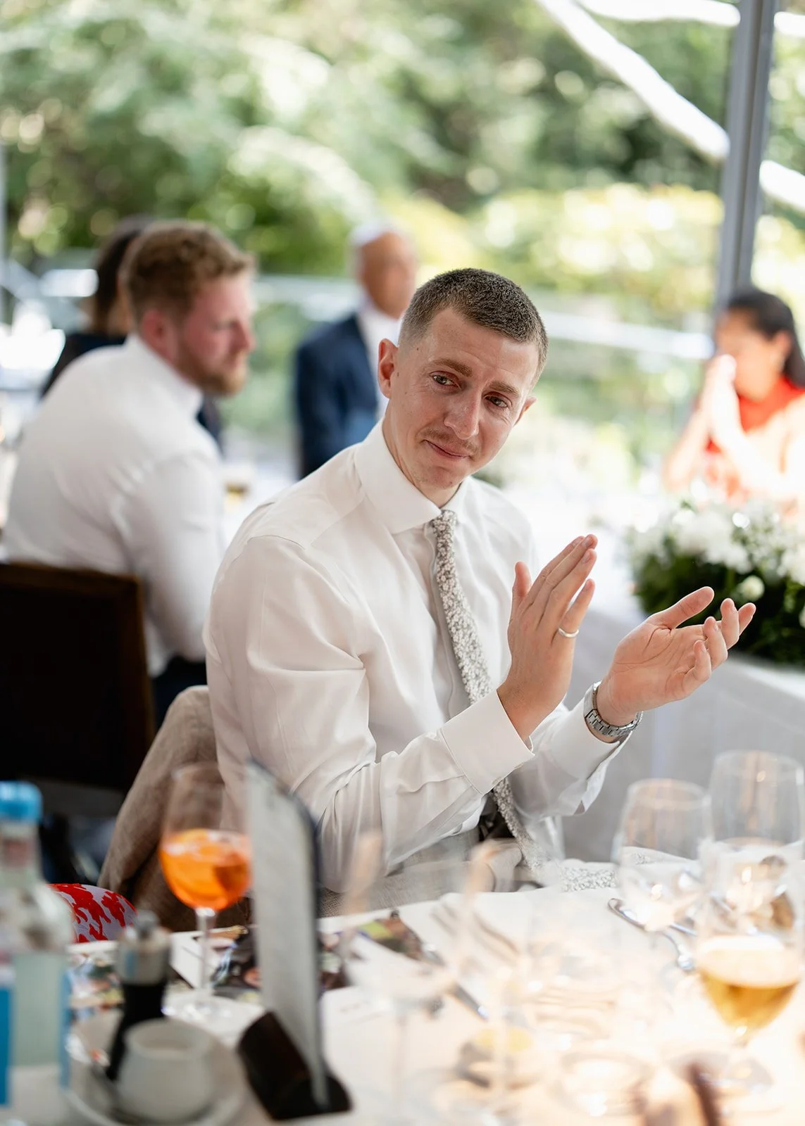 A man in a white shirt and patterned tie raising his hand in a clapping gesture at a table during a formal event, with other people sitting at the table and greenery visible outside through a window.