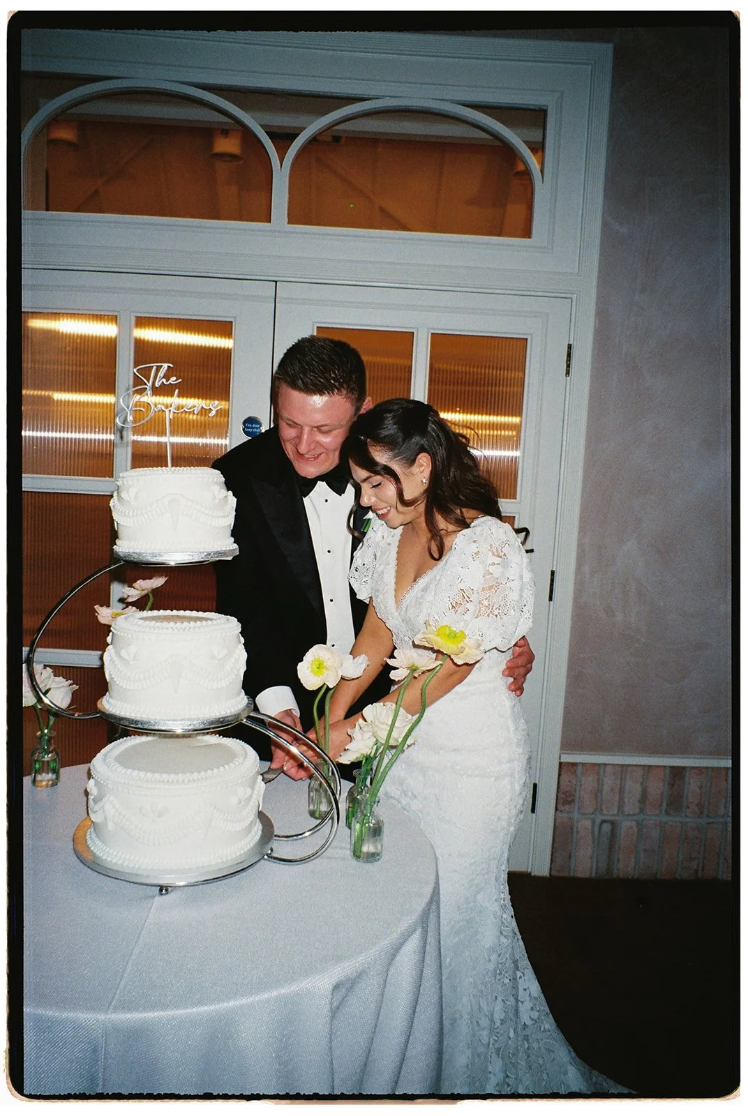 A bride and groom in wedding attire cutting a three-tiered white wedding cake with a gold cake topper that says 'The Goffers.' The bride holds a bouquet of white flowers, and they are both smiling, standing in front of a window with warm lighting.