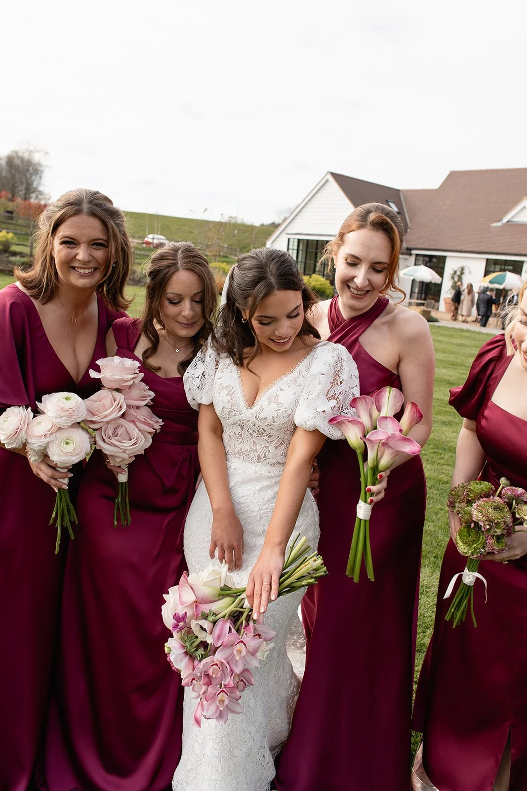 Group of women in formal dresses holding bouquets, including a bride in white and bridesmaids in burgundy, outside on a lawn during a wedding celebration.