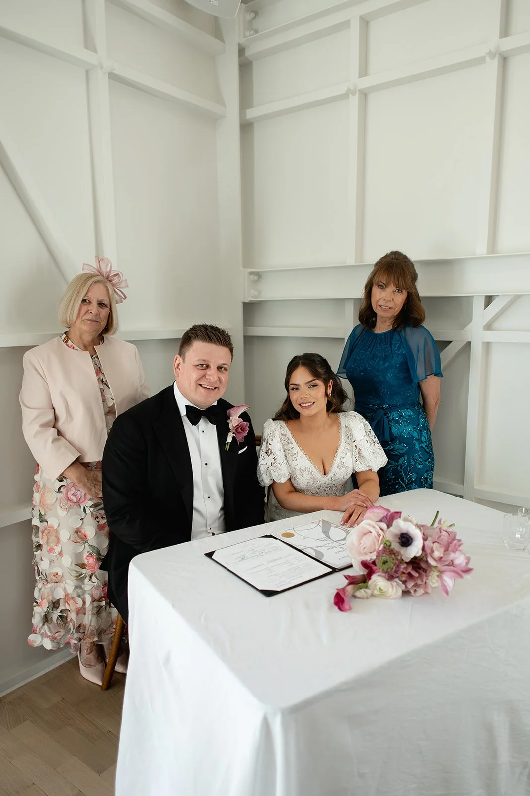 A couple appears to be signing a wedding register at a table, with three women standing behind them, in a bright room with white walls. The couple is dressed in formal wedding attire, with the man in a black tuxedo and the woman in a white lace dress