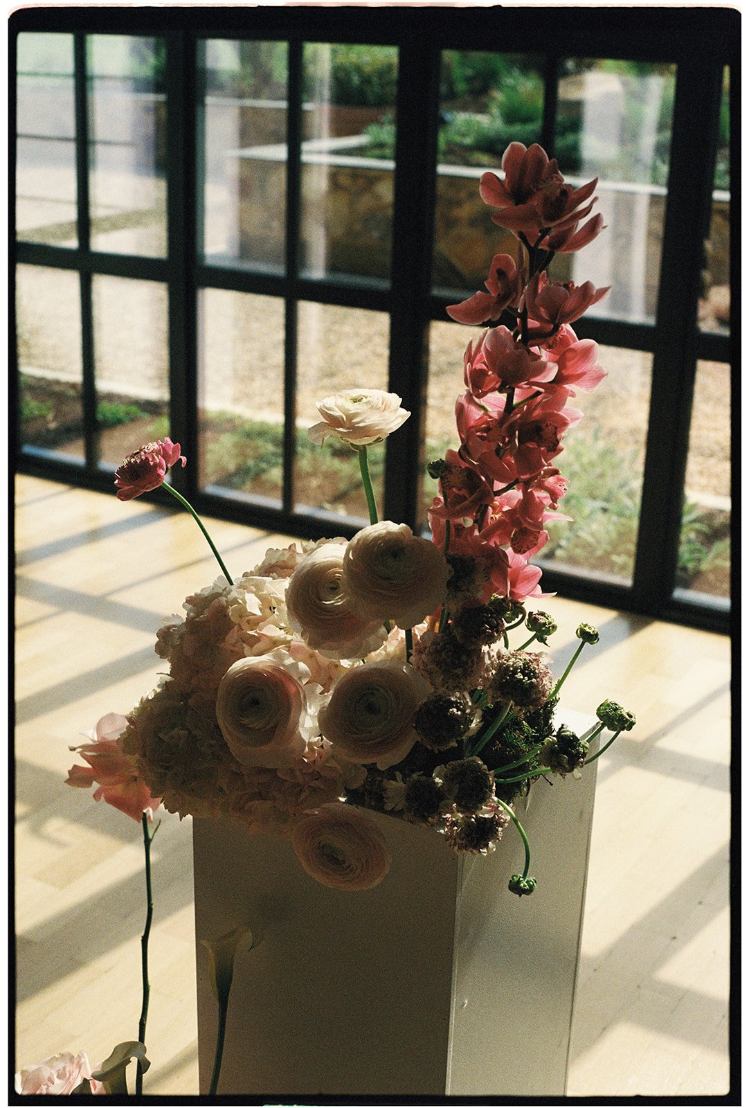 A floral arrangement with pink and white flowers in a white vase, set on a table near sliding glass doors with a garden view.