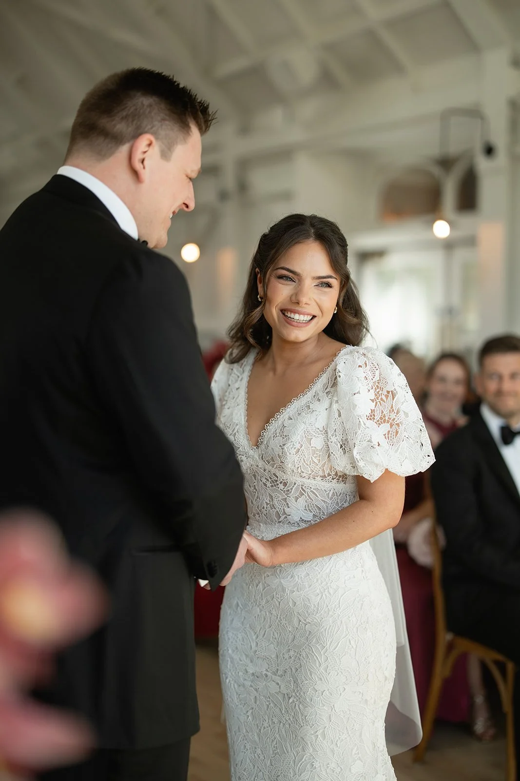 A bride and groom during their wedding ceremony, with the bride smiling and looking at the groom who is holding her hands.