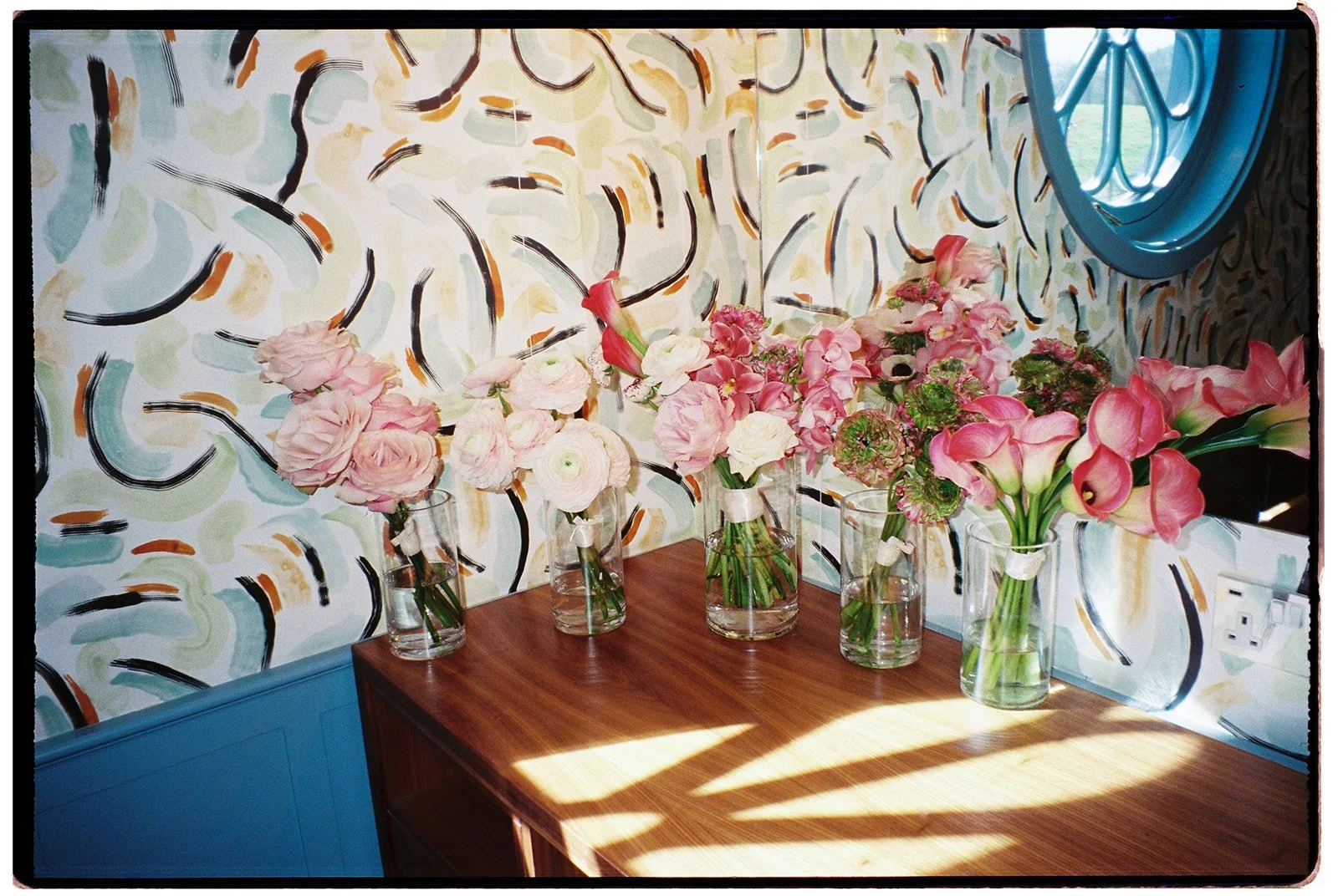 Five glass vases with pink and white flowers arranged on a wooden table in front of a colorful abstract patterned wall and a mirror.