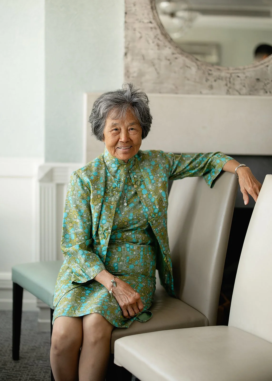 An elderly woman with gray hair, smiling, sitting on a beige chair in a room with neutral decor, wearing a patterned green and blue outfit and accessories.