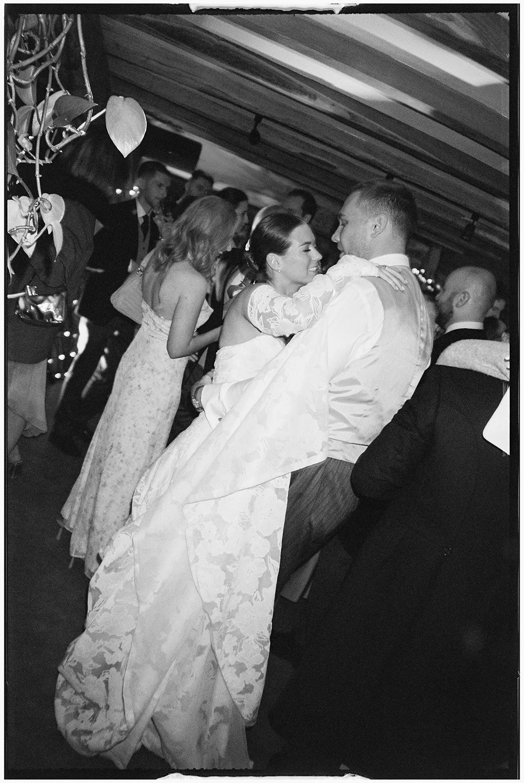 A bride and groom dancing closely at a wedding reception, surrounded by guests, some of whom are dancing or talking in a decorated indoor venue.