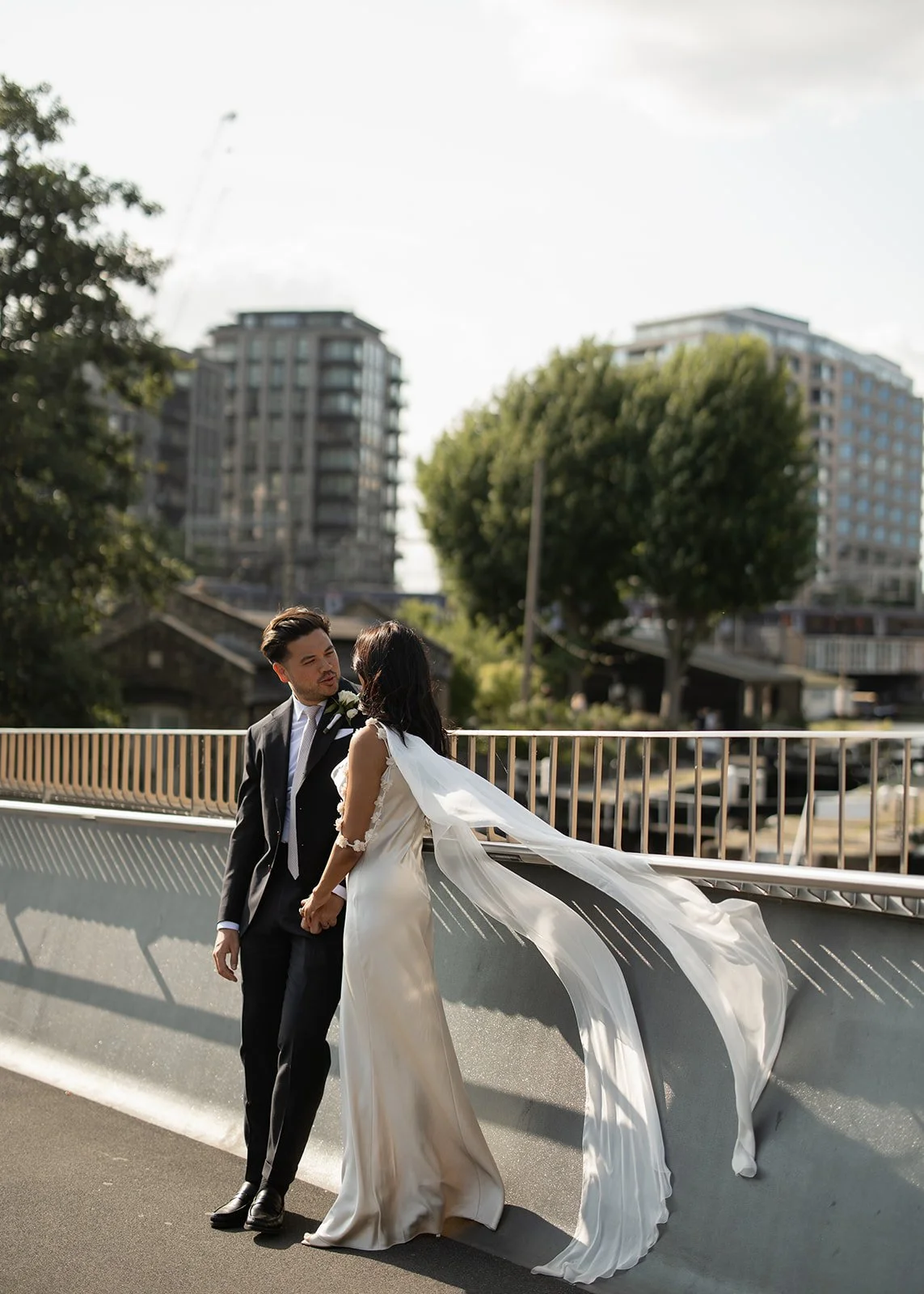 A couple dressed in wedding attire standing on a bridge holding hands, with the bride's long veil floating in the wind. Urban buildings and trees are in the background on a sunny day.