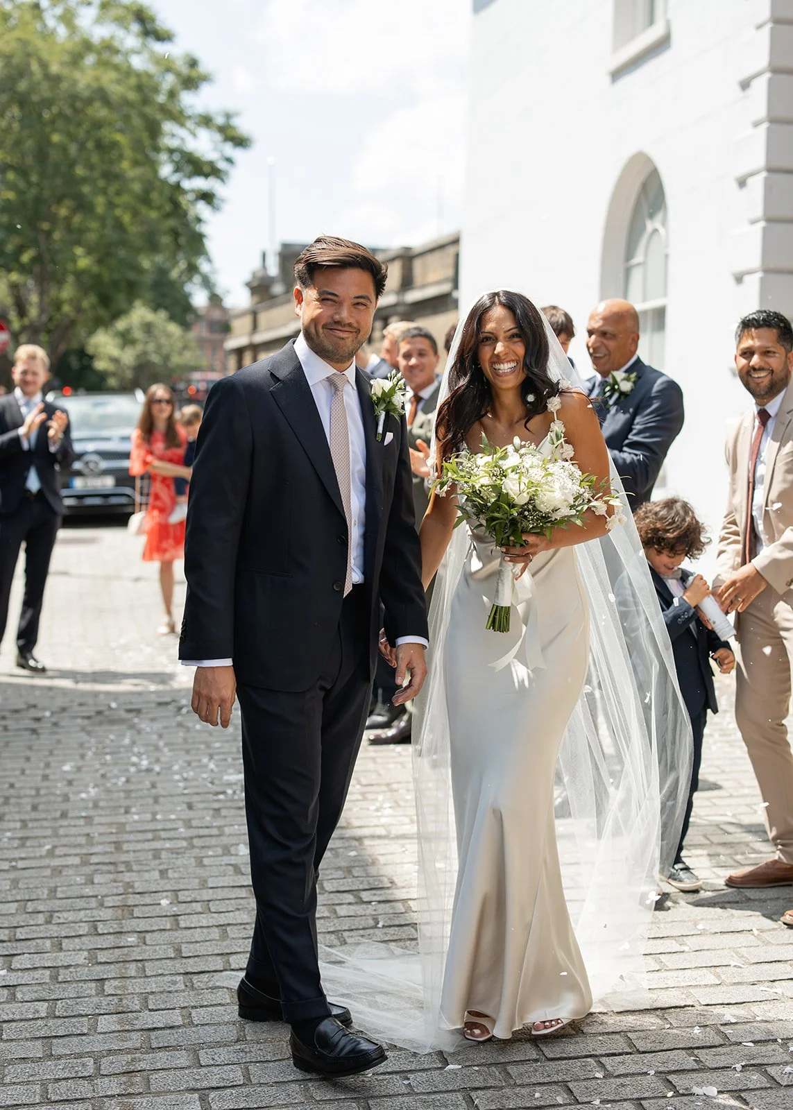 A newly married couple walking outdoors on a cobblestone street, holding hands and smiling. The bride is wearing a white wedding dress with a bouquet of flowers, and the groom is dressed in a black suit. Several guests are celebrating behind them, clapping and smiling.