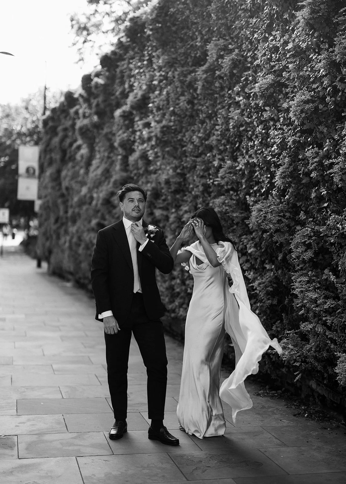 Black and white photo of a man in a suit and a woman in a flowing gown walking along a sidewalk next to a hedge, with the woman covering her face with her hands.