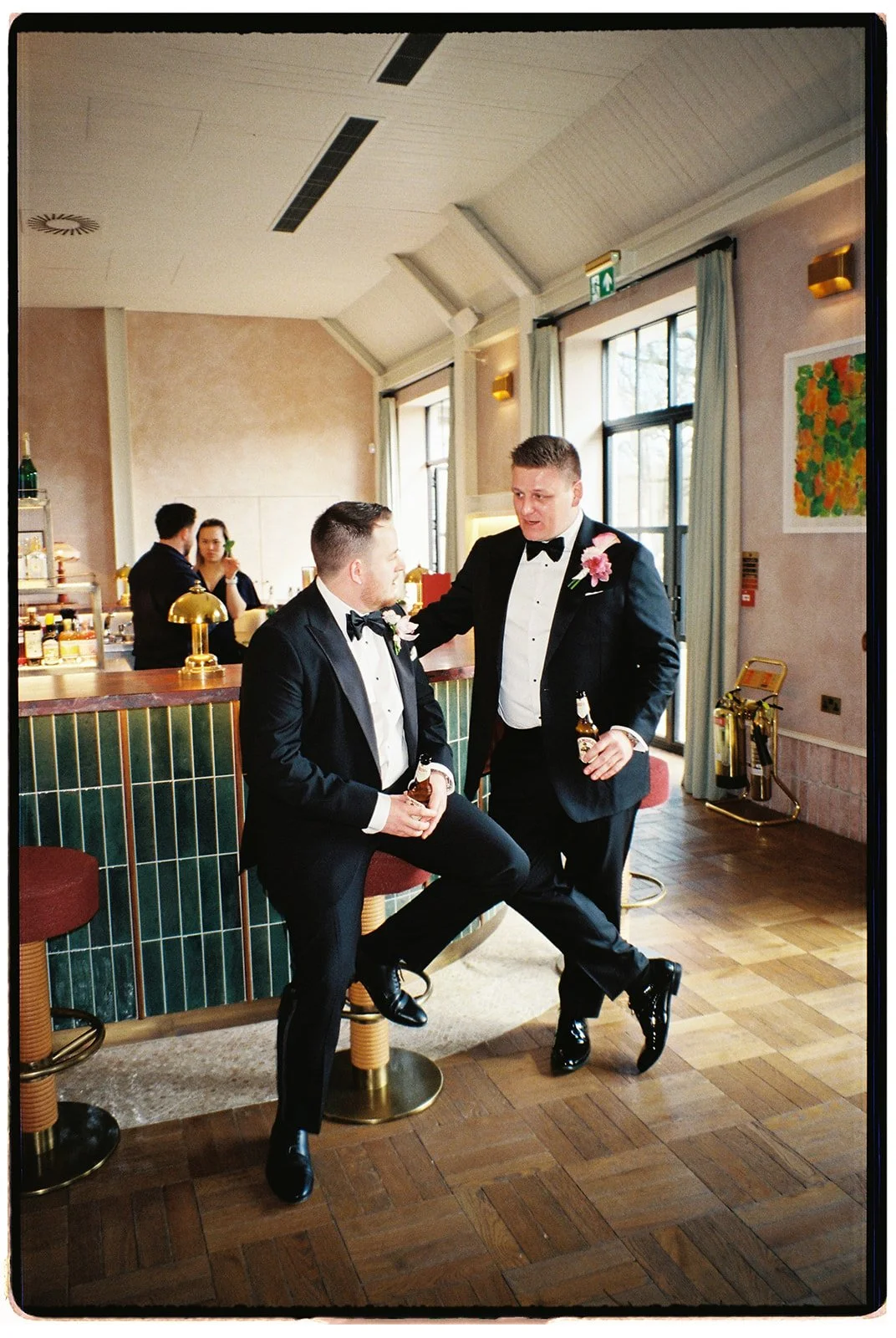 Two men in tuxedos with pink boutonnieres are having a conversation at a bar, holding drinks, in a well-lit room with large windows, curtains, and colorful artwork on the wall.