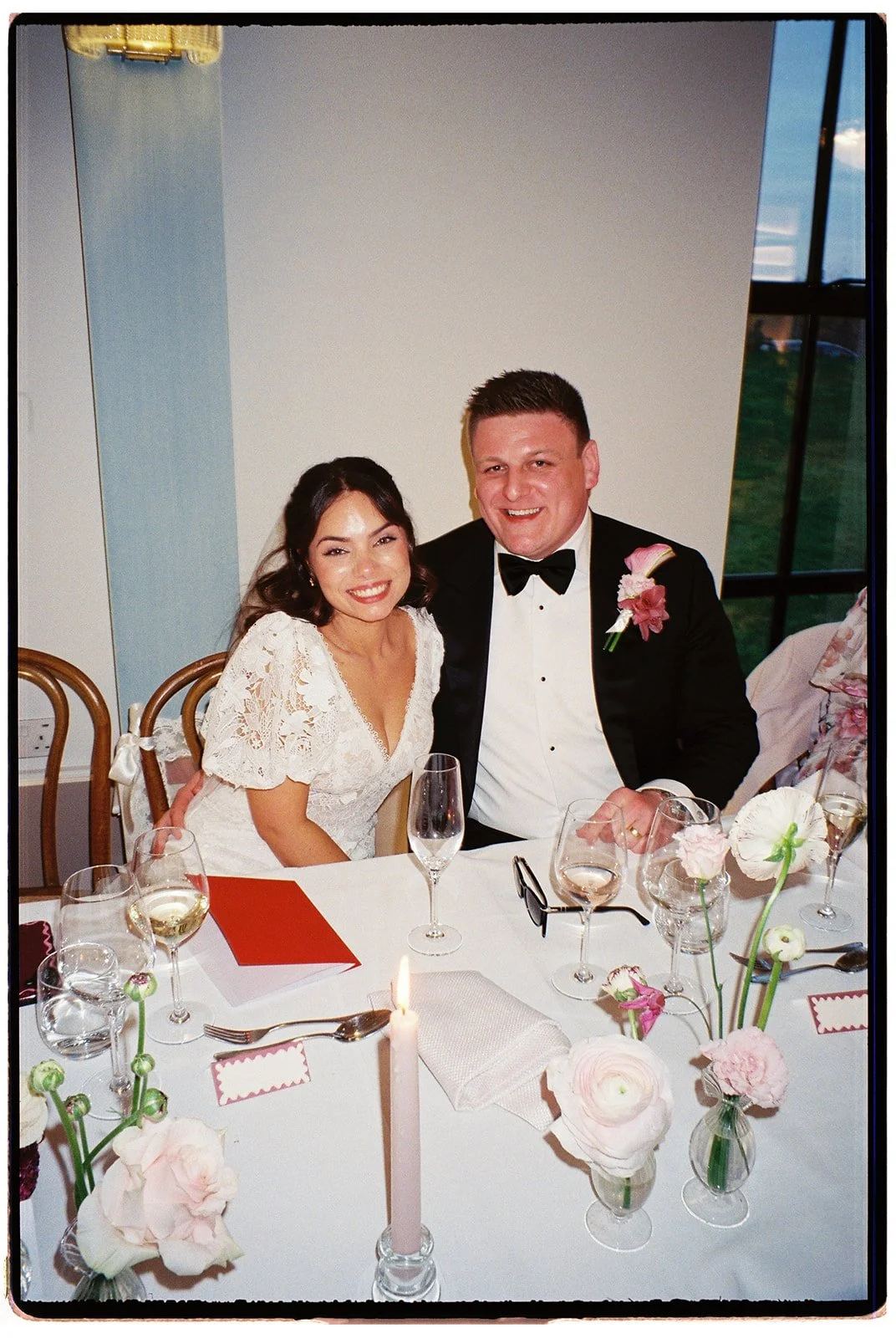 A newlywed couple sitting at a decorated wedding reception table, smiling, with flower arrangements and glasses of wine in front of them.