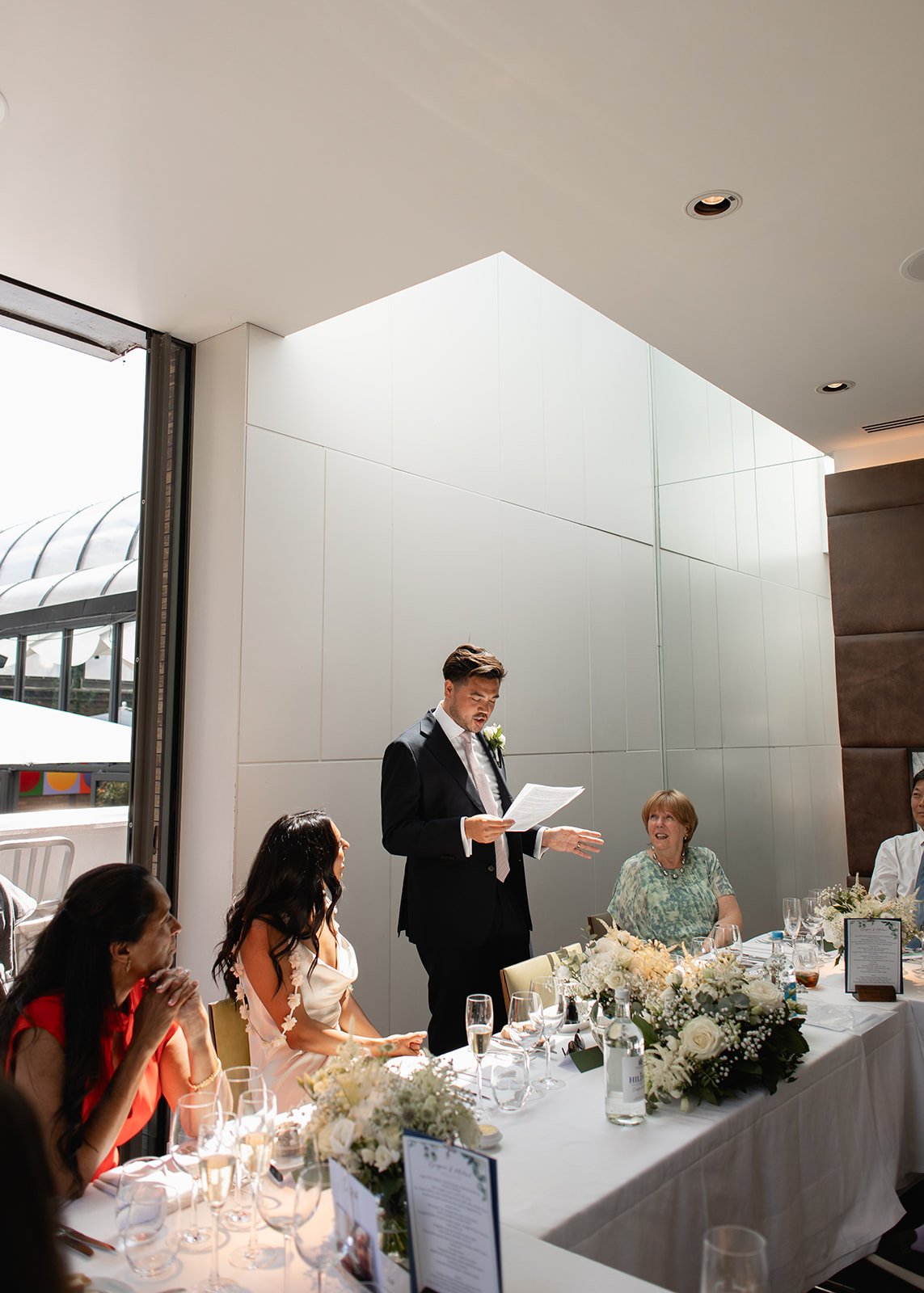 Man in a black suit giving a speech at a wedding reception, seated at a decorated table with women listening, in a bright, modern room.
